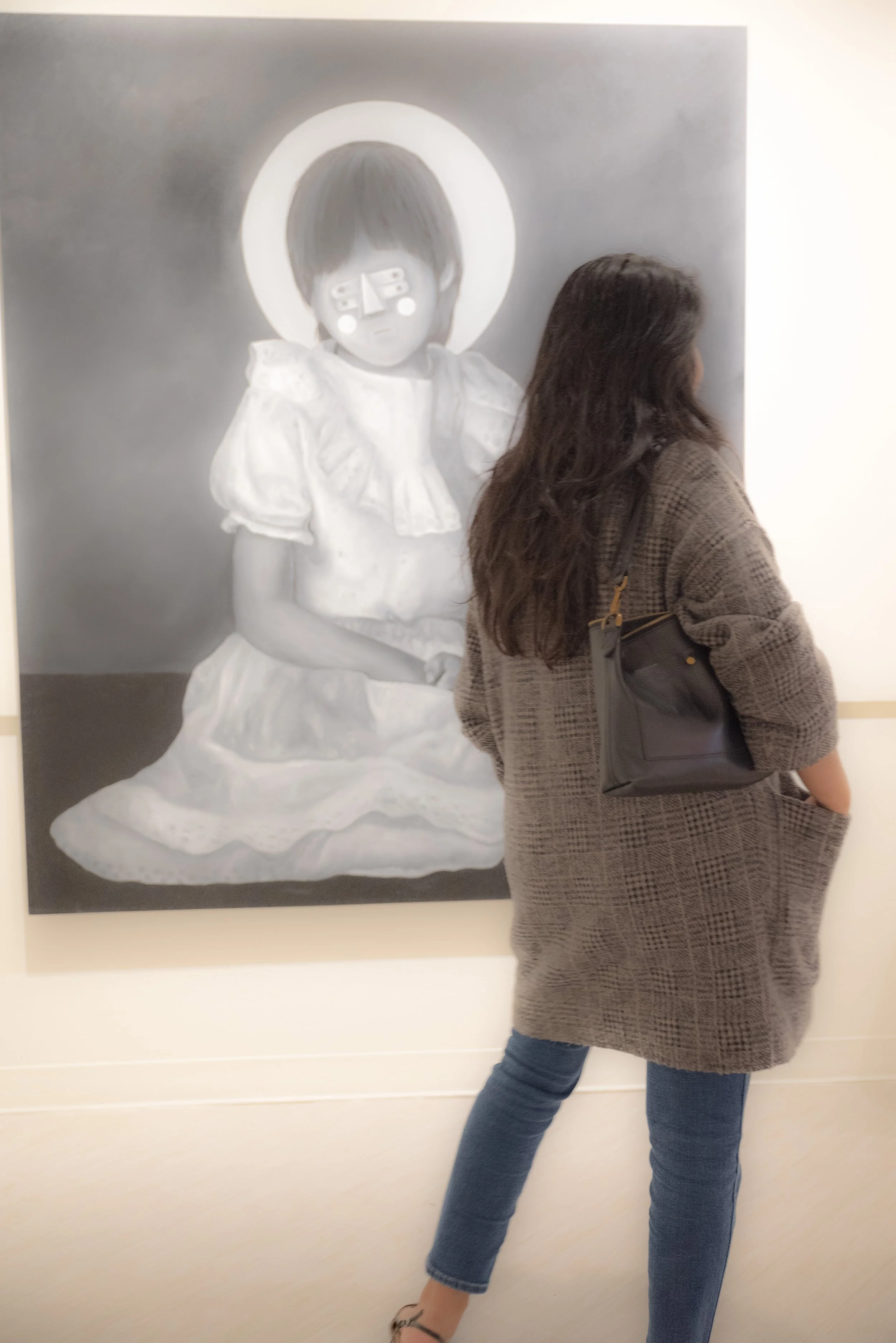 A woman with long dark hair wearing a gray coat and carrying a black handbag is standing in front of a large black-and-white painting of a girl with short hair, wearing a dress with a ruffled collar, and sitting on the ground with a halo around her h