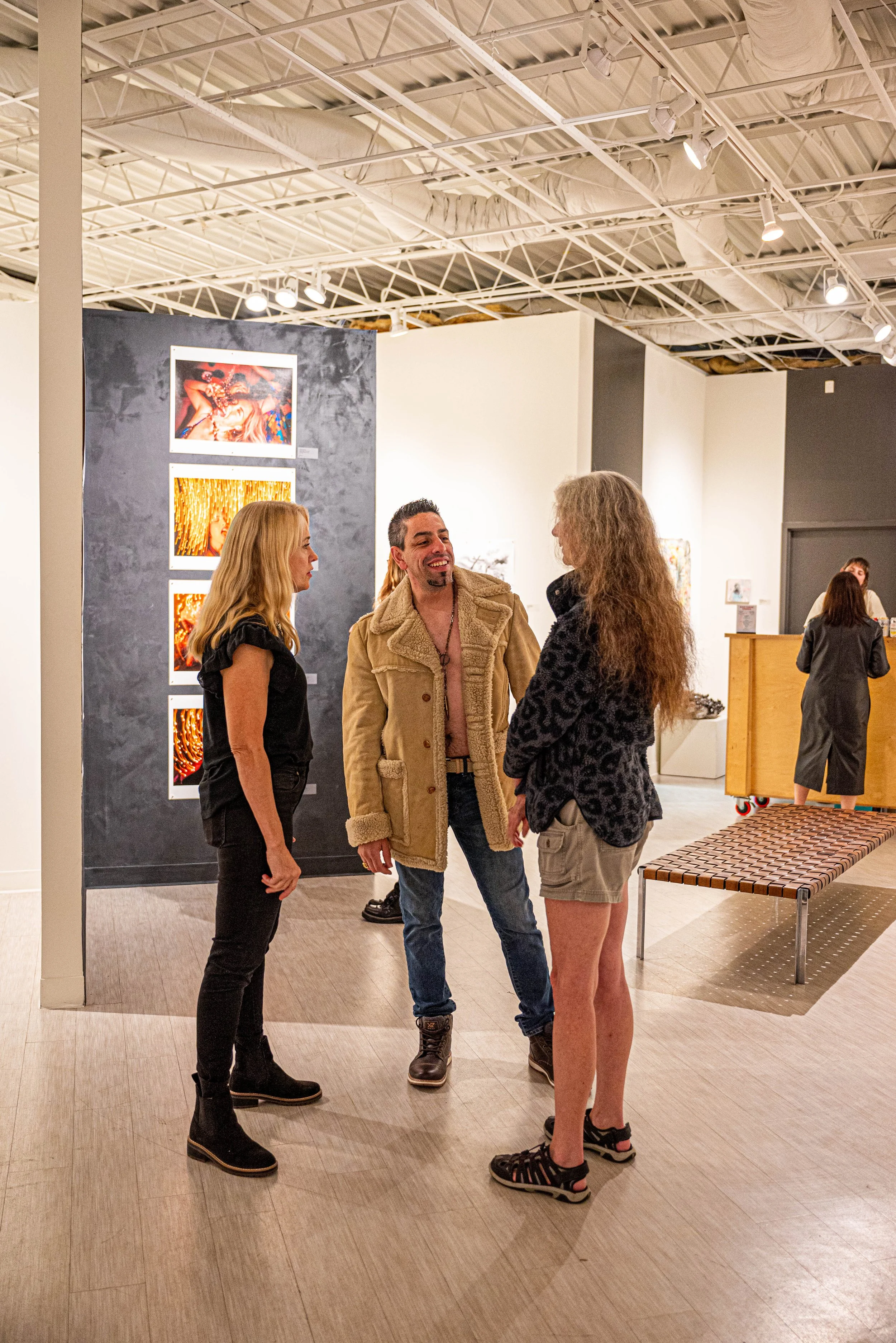 Three people are standing and talking in an art gallery with artworks on the wall behind them. One person is smiling, and the other two are listening. There are people at a counter in the background.