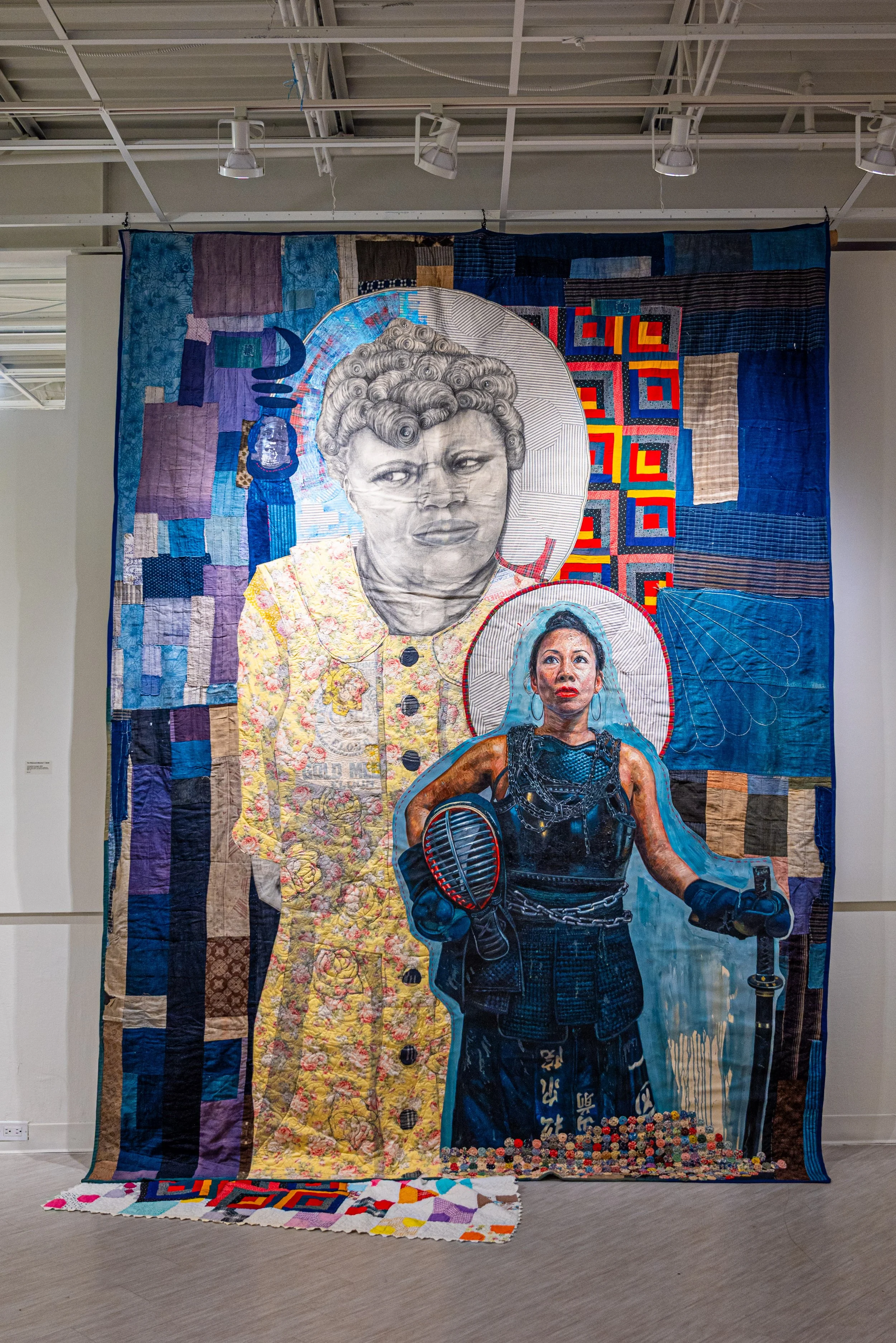 A large textile artwork featuring two women with an abstract patchwork background. The woman in the foreground is dressed as a fighter with boxing gloves and a chain, and the woman in the background is depicted in a black-and-white portrait style.