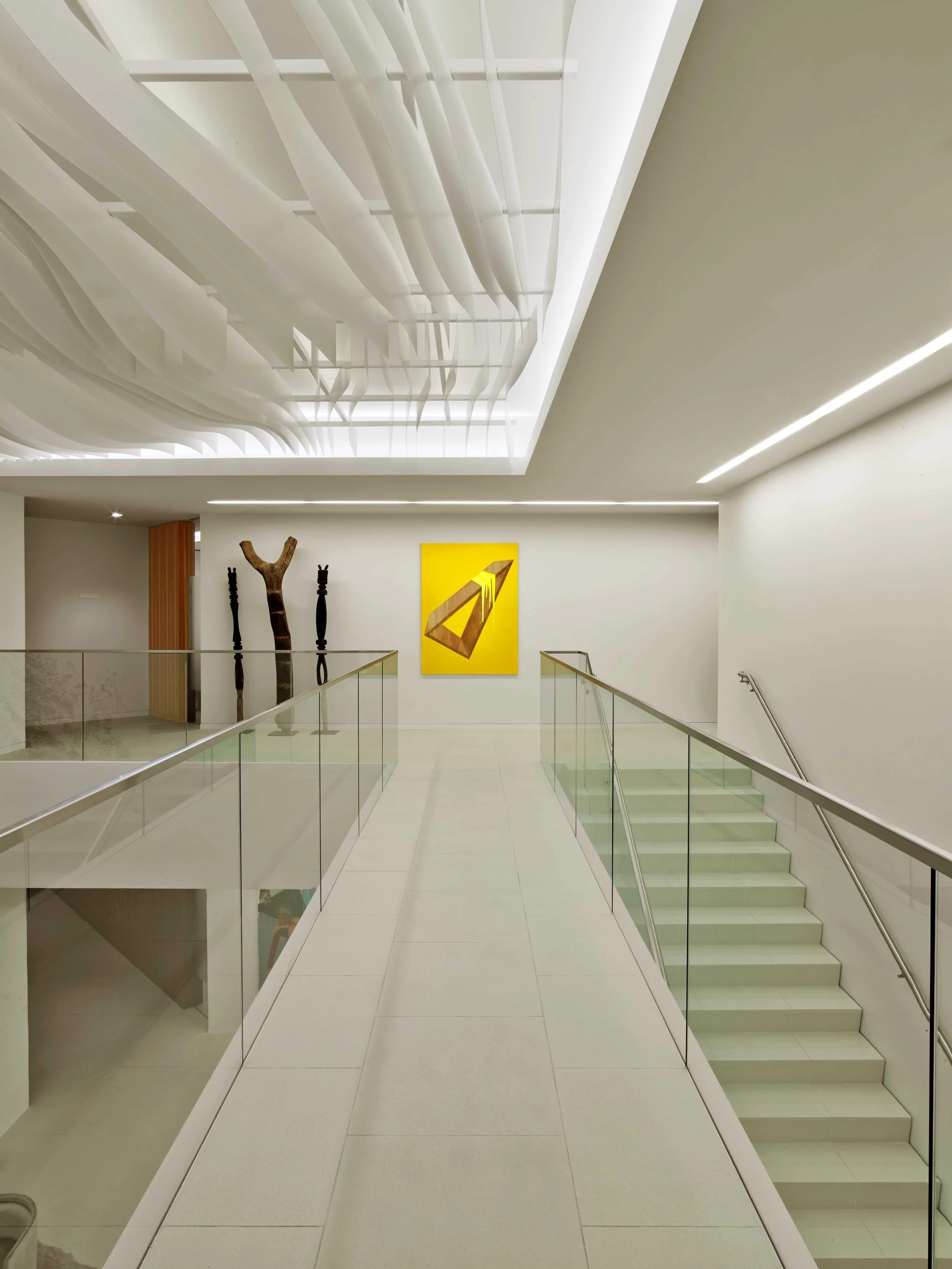 Modern art gallery interior with staircase, abstract yellow painting, wooden sculptures, and a ceiling installation of white curved panels.