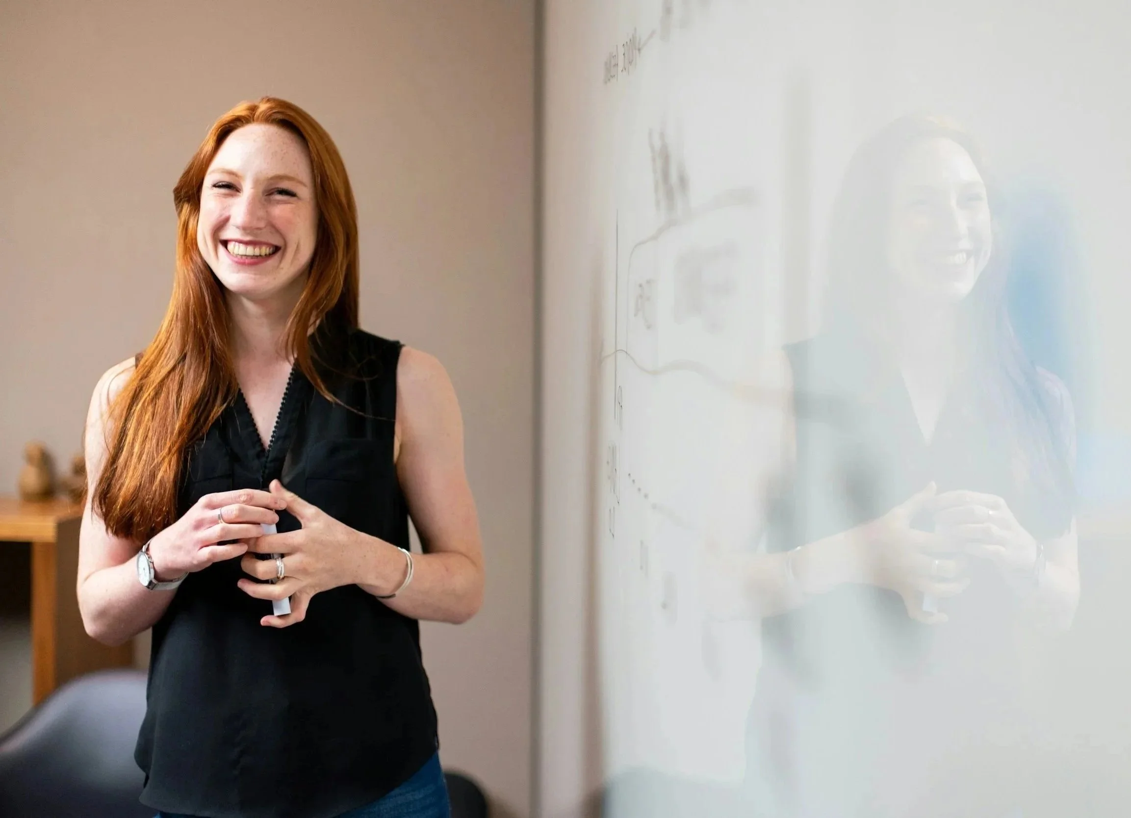 A smiling woman with long red hair in a black sleeveless top standing in front of a whiteboard with her reflection visible on it.