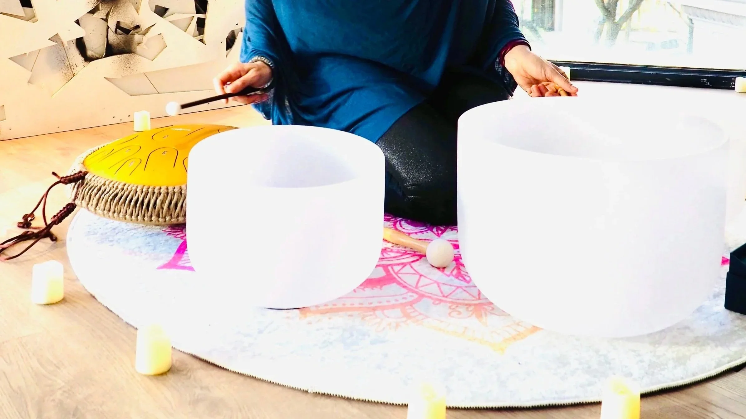 Person playing wind chimes and singing bowls during a sound healing session, with a candle-lit circle on the floor.