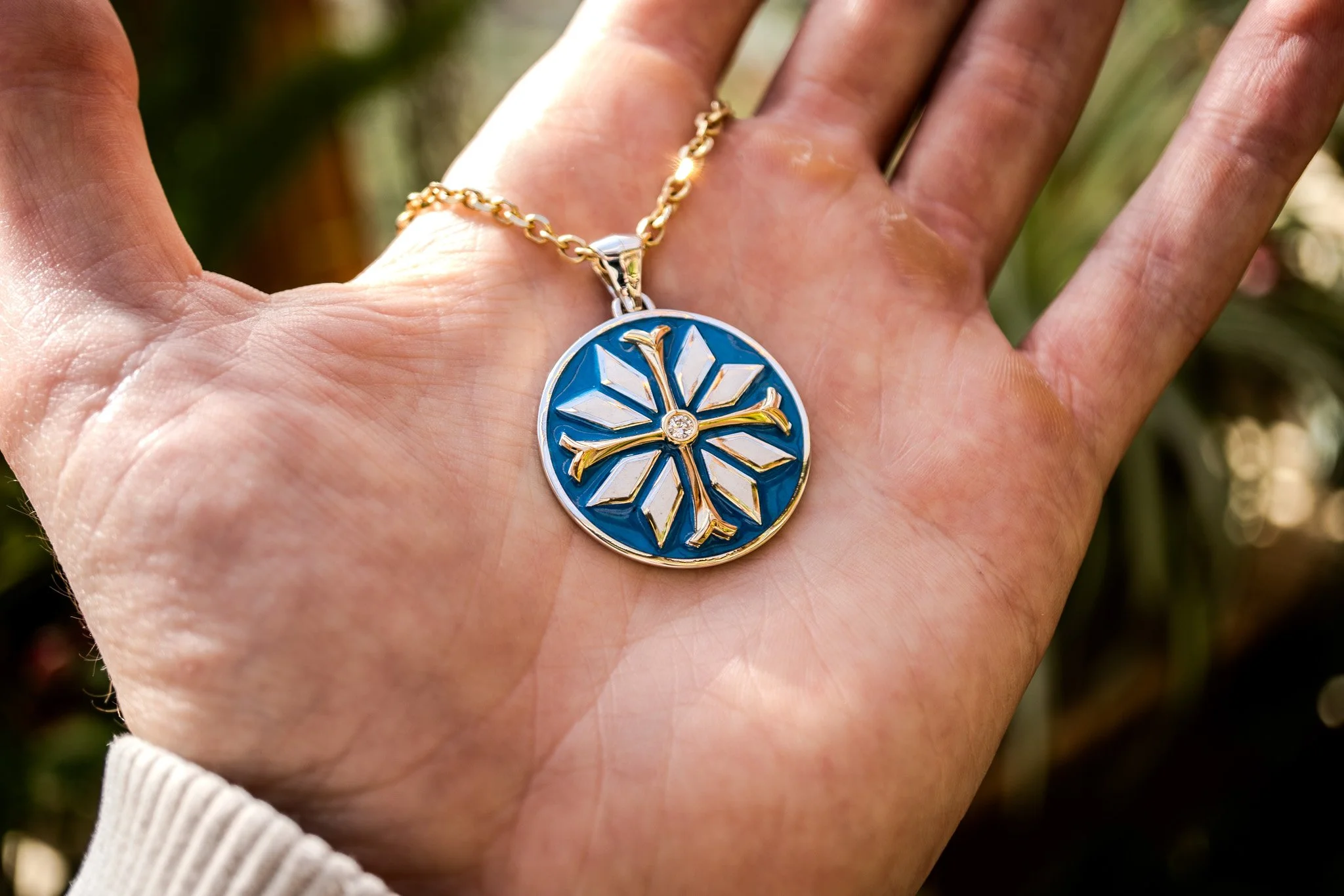A person's hand is holding a gold chain necklace with a round pendant. The pendant has a blue background with a white snowflake design and gold accents, with a small gemstone in the center.