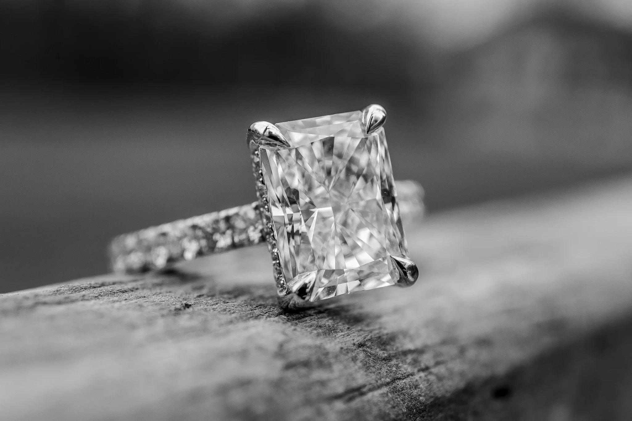 Close-up of a large, rectangular-cut diamond ring with smaller diamonds along the band, resting on a textured surface. Black and white photography.