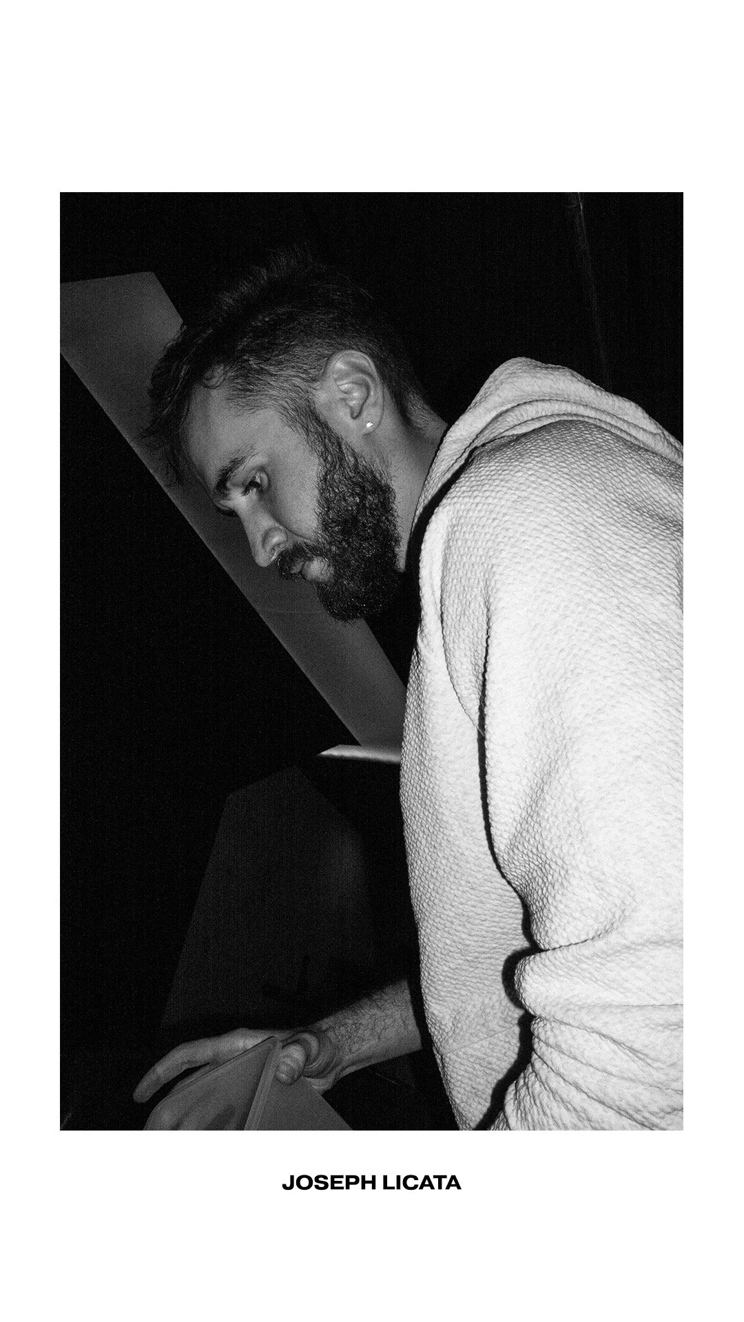 Black and white portrait of a man with a beard, earring, and textured hoodie, looking down, with the name Joseph Licata beneath the image.