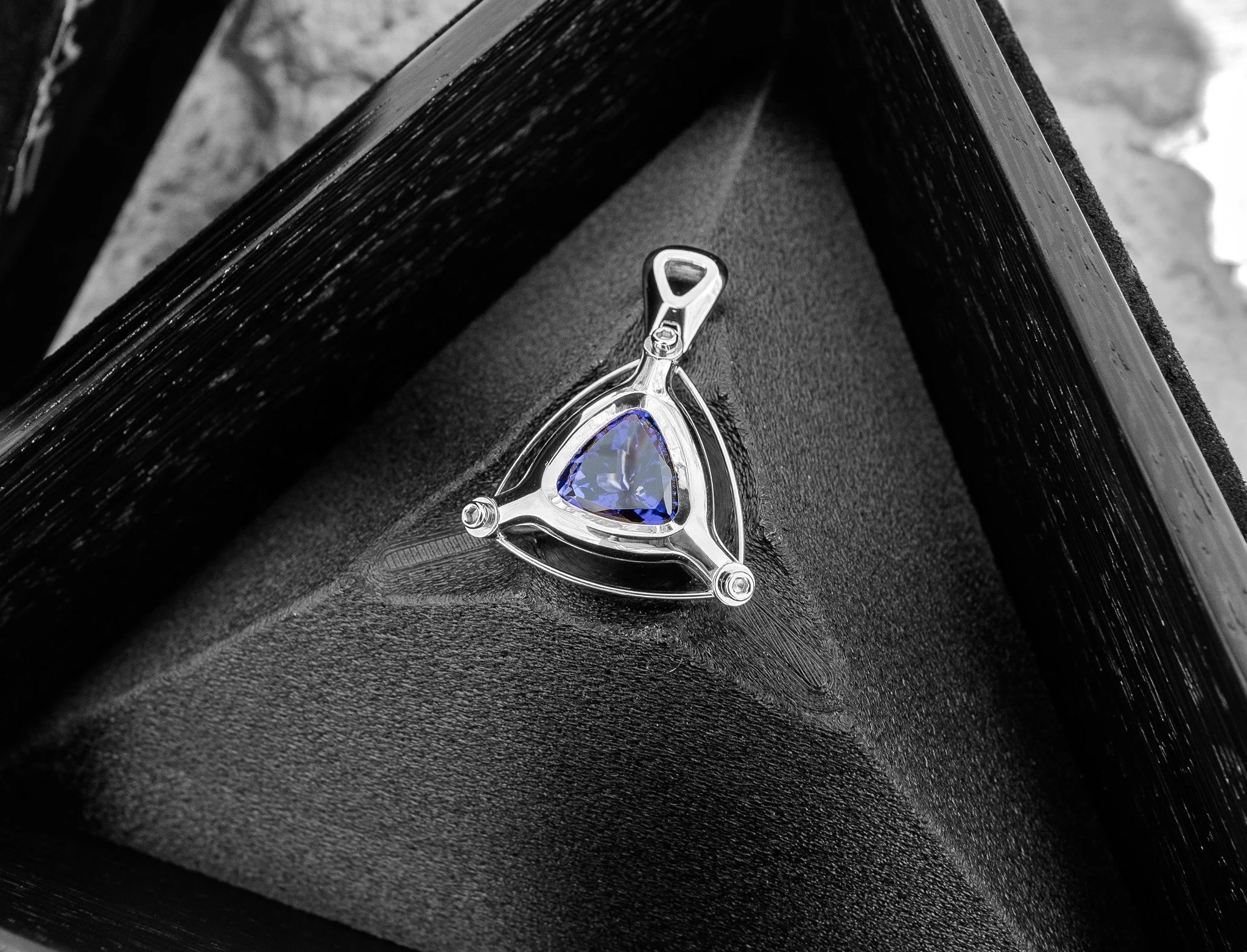 A silver pendant with a triangular blue gemstone resting inside a black jewelry box.