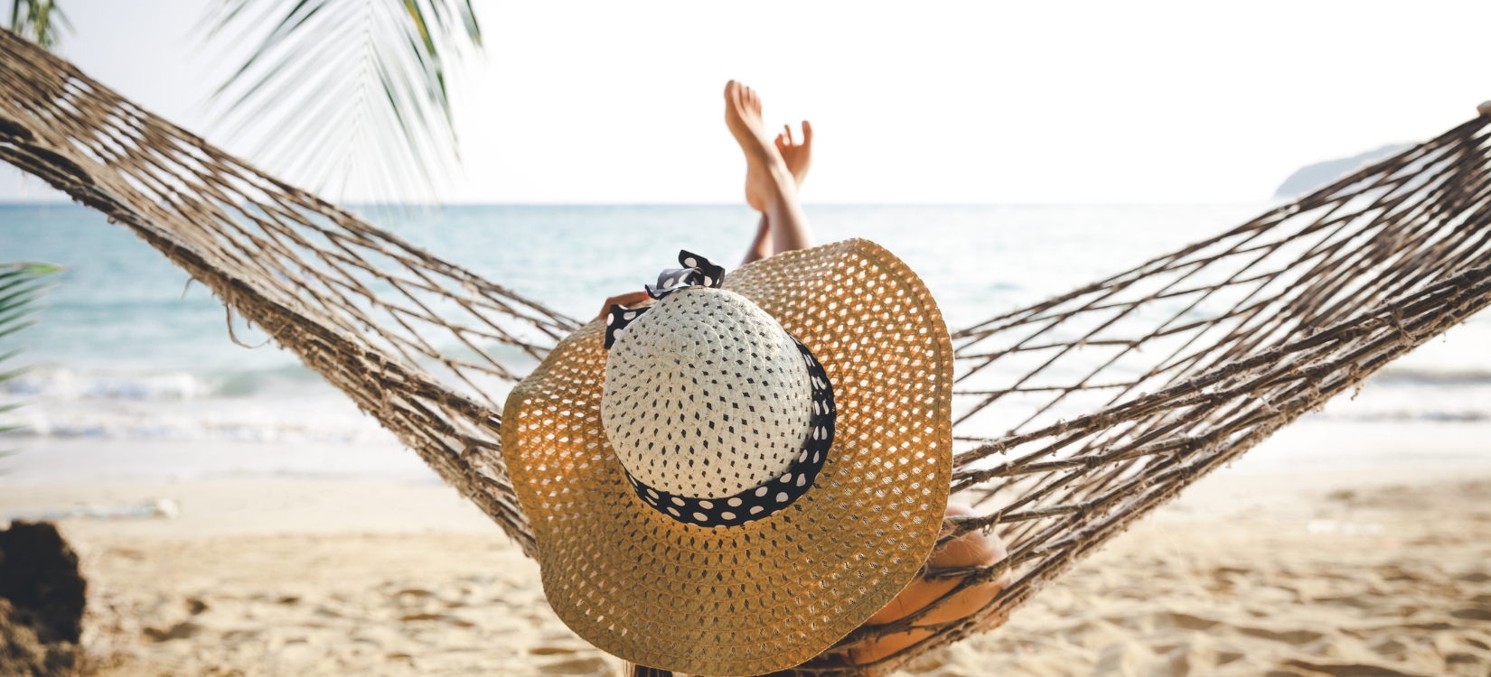 a woman is on the beach, in a hammock. we can only see her from behind obscured by a large hat