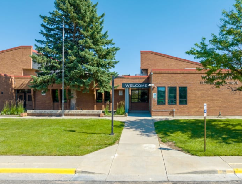 Front entrance of a brick school building with a welcoming sign and an American flag pole, green lawn, and trees.