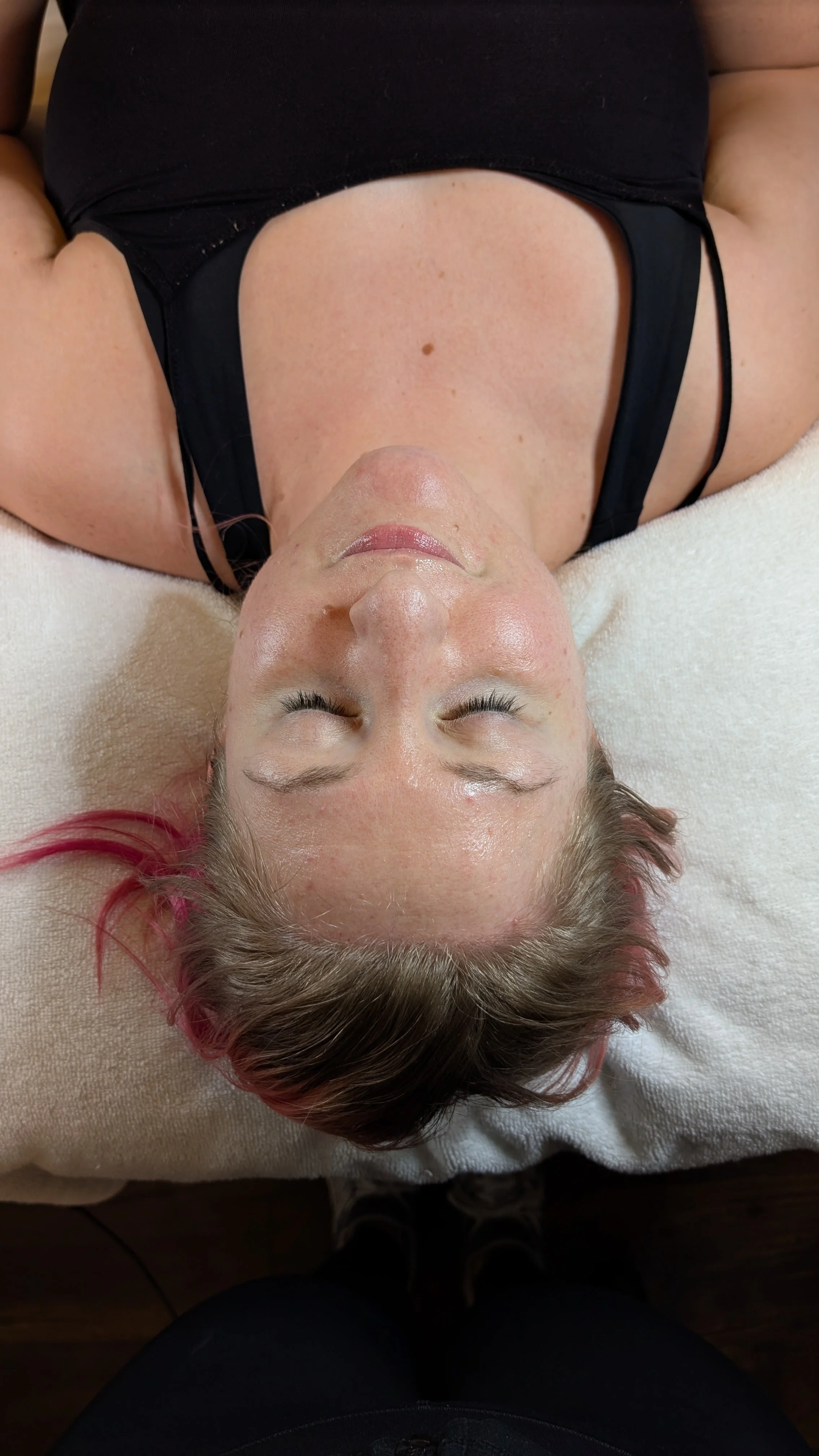A woman with pink-highlighted hair lying on her back on a white surface, eyes closed, wearing a black top.