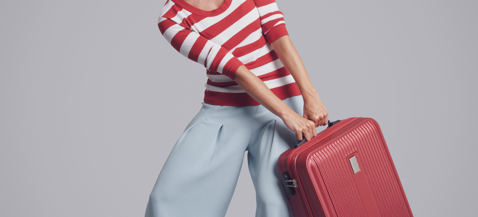 a woman is carrying luggage. she is wearing a striped red shirt and wide leg trousers. she is holding the bag extremely off centered