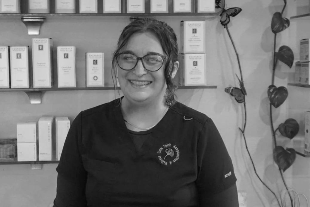 Black-and-white photo of a smiling woman with long dark hair wearing a dark scrub top, standing in front of shelves with skincare or health products Ariel Donofrio