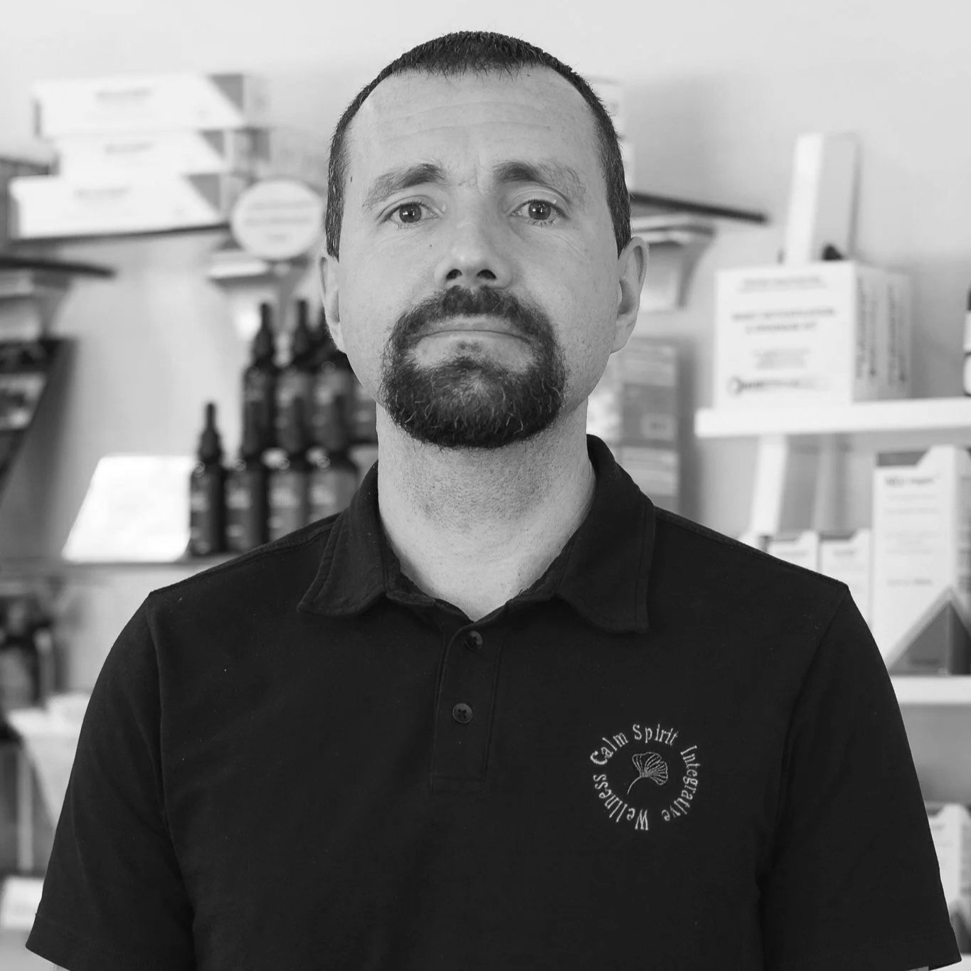 A man with a beard and mustache standing outdoors near trees with a cloudy sky in the background, wearing a hoodie with hands in pockets. Black and white photograph Andrew MacNeil
