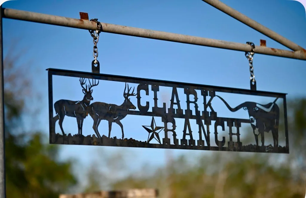 Metal sign with the text 'Clark Ranch' hanging from a horizontal bar, featuring silhouettes of three deer, a star, and a longhorn steer, against a blurred outdoor background with blue sky.
