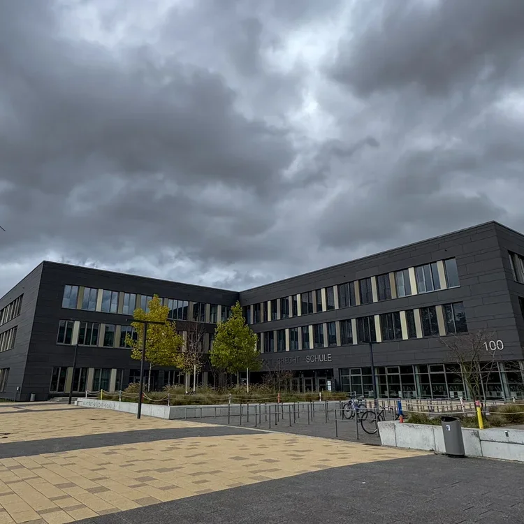 Neubau der Bertolt-Brecht-Schule in Nürnberg: Modernes dreigeschossiges  Schulgebäude mit schwarzer Fassade und großen Fenstern, davor gepflasterter  Platz mit jungen Bäumen, dramatisch bewölkter Himmel