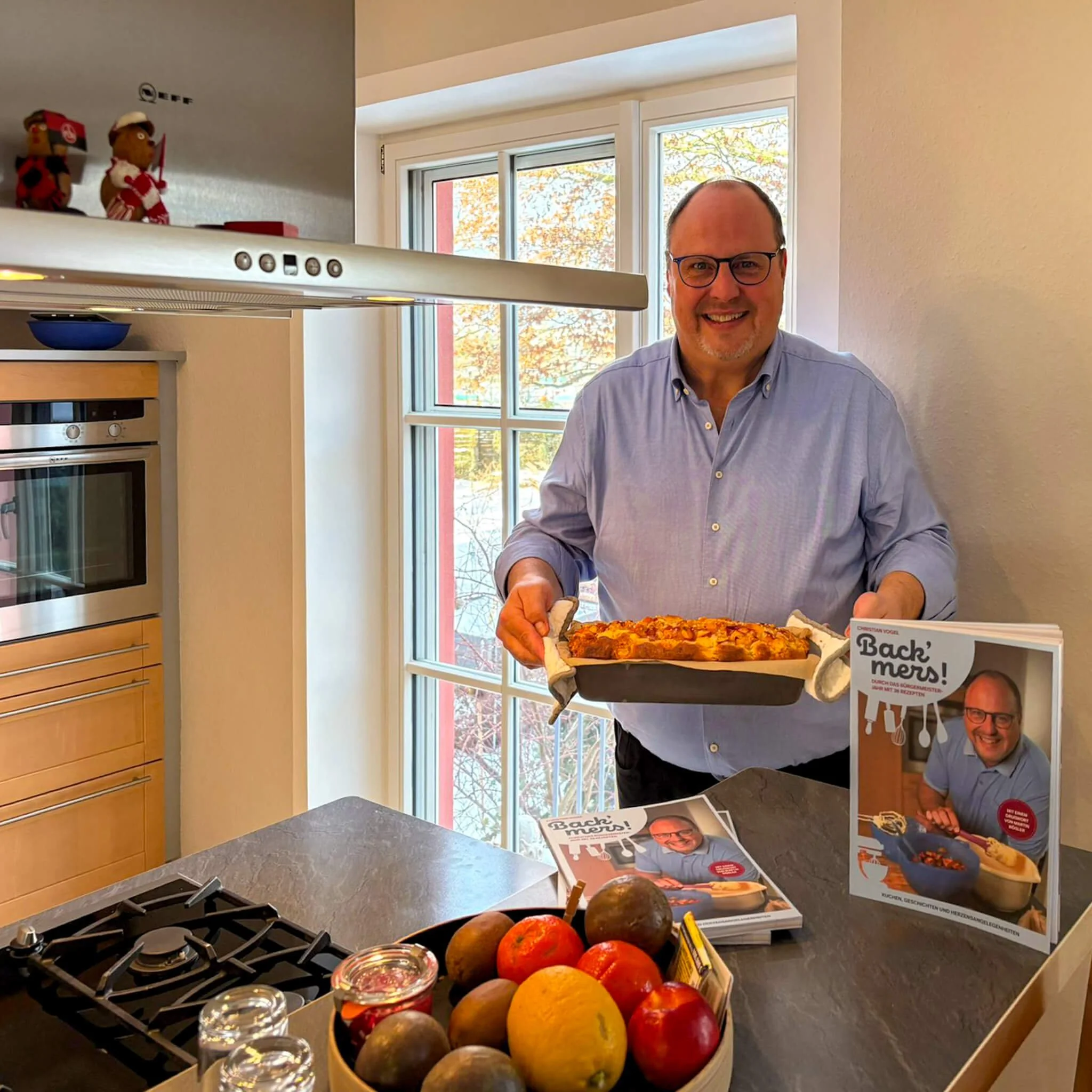 Nürnbergs Bürgermeister Christian Vogel lächelt in einer Küche, mit selbstgebackener Torte und seinem Backbuch "Back' mers!"