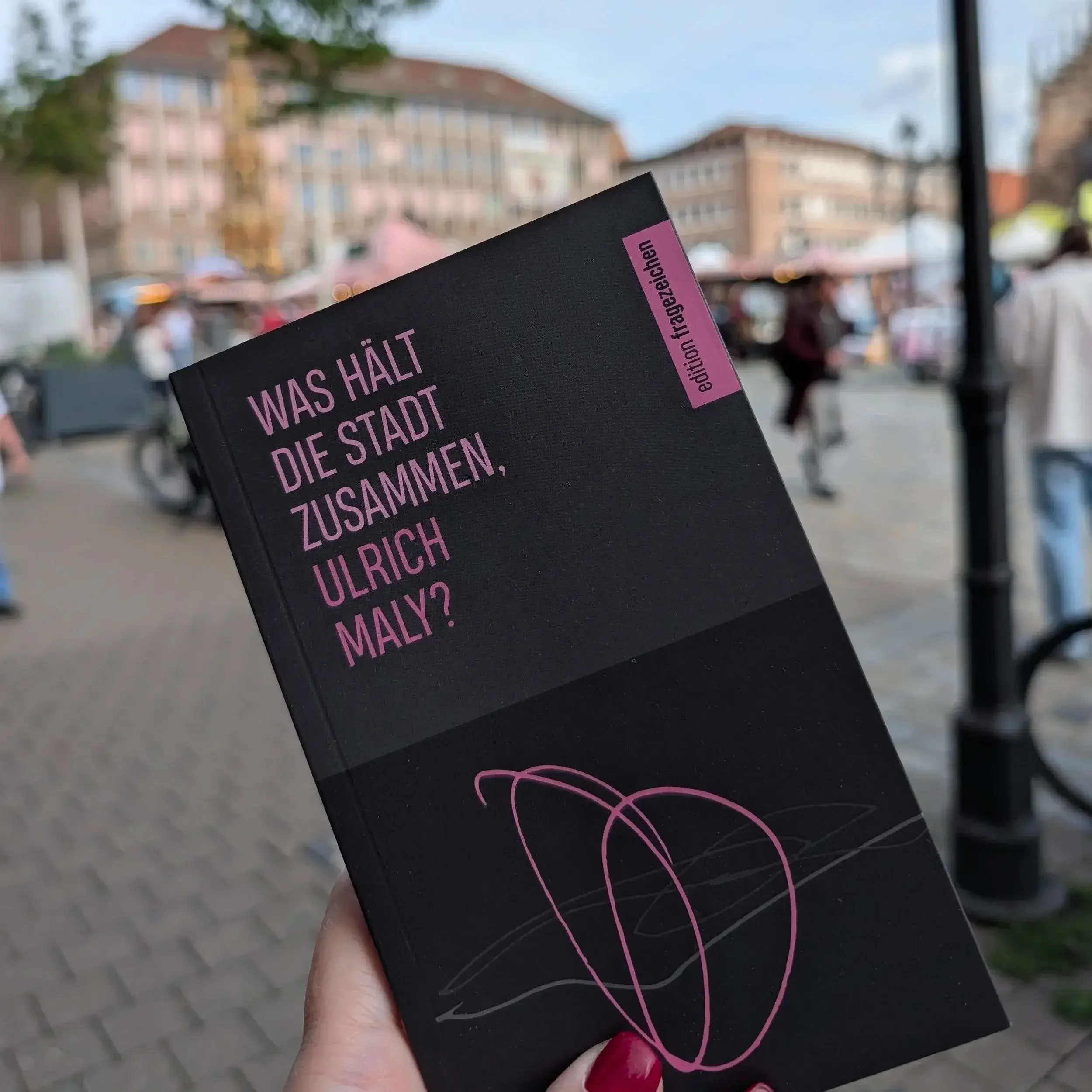 Eine Hand hält Ulrich Malys Buch „Was hält die Stadt zusammen, Ulrich Maly?“ vor dem Hauptmarkt in Nürnberg.