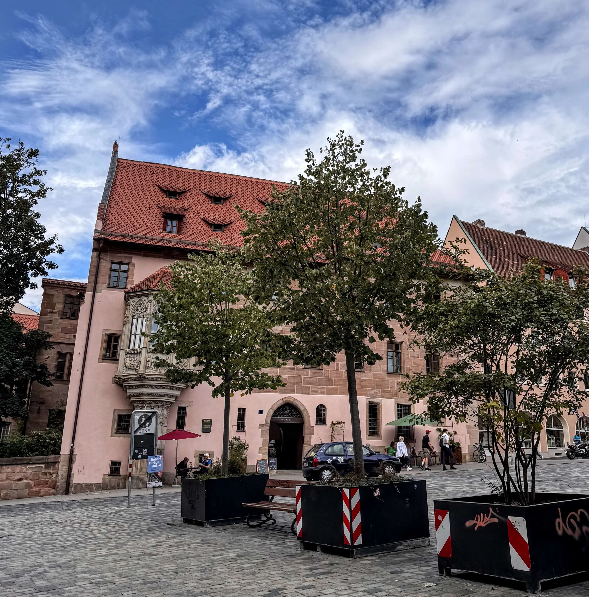 Bäume in schwarzen Pflanzkübeln mit rot-weißen Warnmarkierungen vor einer historischen Fassade St. Sebald mit steinernem Erker in Nürnberg am Sebaldusplatz.