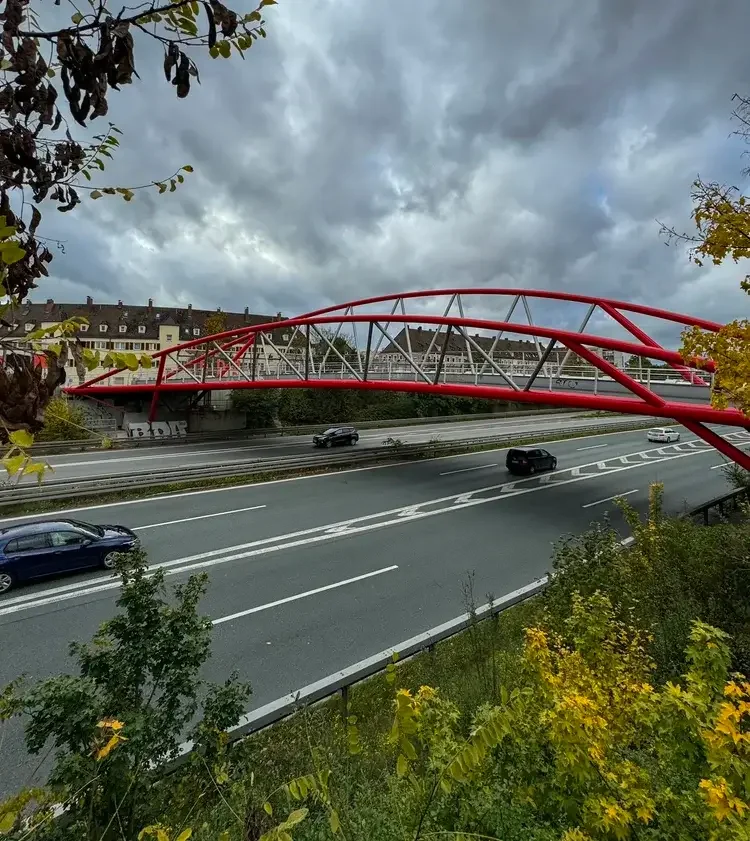 Rote Stahlbogenbrücke überspannt den Frankenschnellweg in Gibitzenhof, im Hintergrund Wohngebäude, dramatisch  bewölkter Himmel, herbstliche Vegetation im Vordergrund