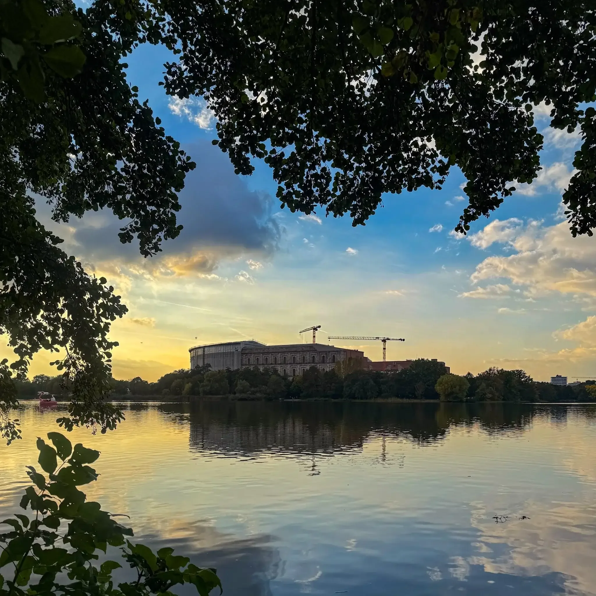 Blick über den Dutzendteich auf die Nürnberger Kongresshalle mit Baukränen bei Sonnenuntergang, gerahmt von Bäumen.
