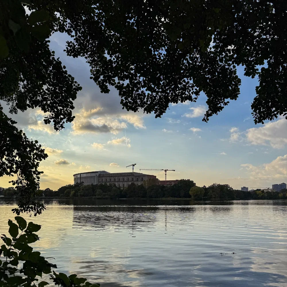 Blick durch Bäume über den Dutzendteich auf die Kongresshalle in Nürnberg mit Baukränen im Hintergrund unter bewölktem Himmel.