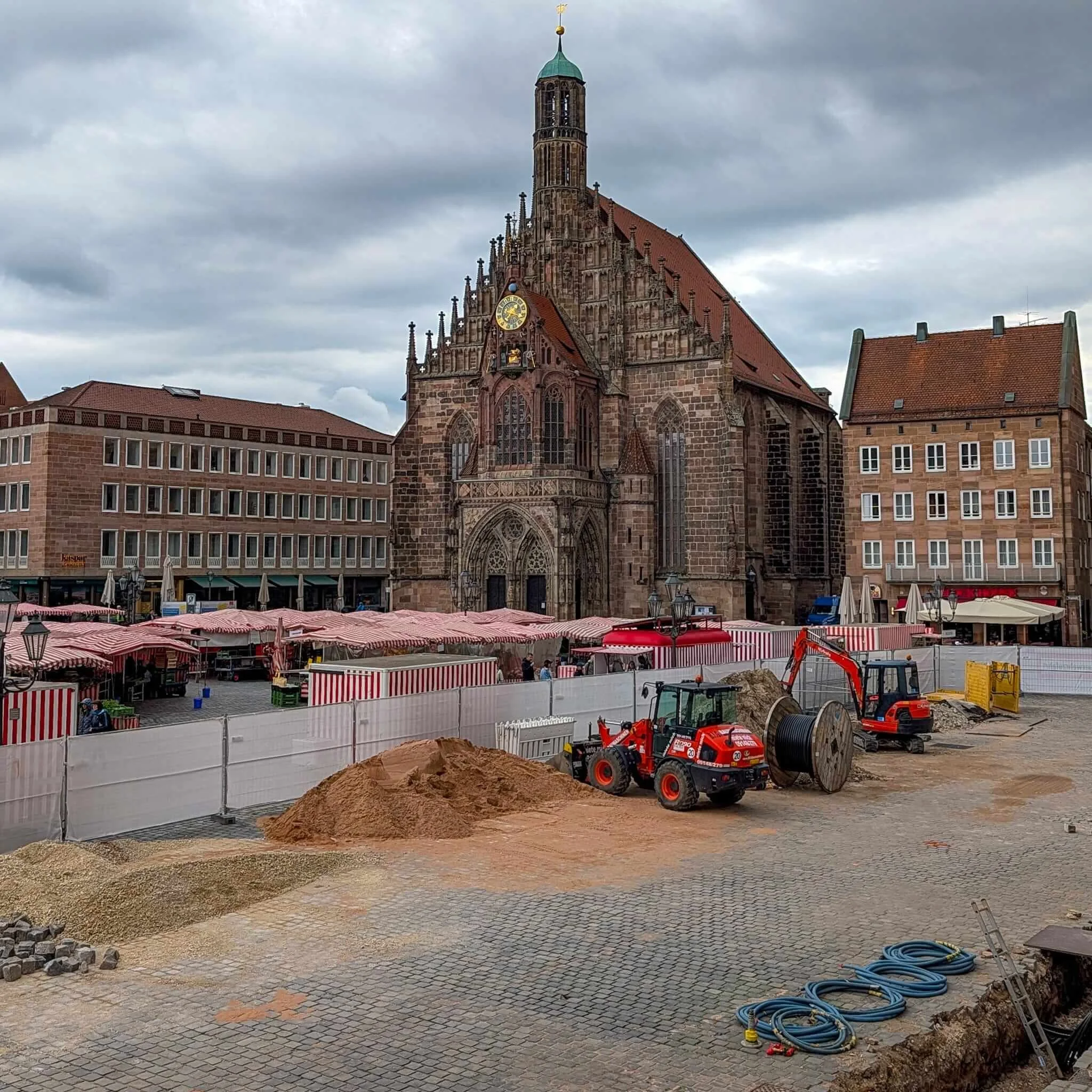 Großbaustelle mit Baggern, Sandhaufen und Bauzäunen auf dem Nürnberger Hauptmarkt. Dahinter stehen die rot-weißen Marktstände des Grünen Markts vor der historischen Frauenkirche.