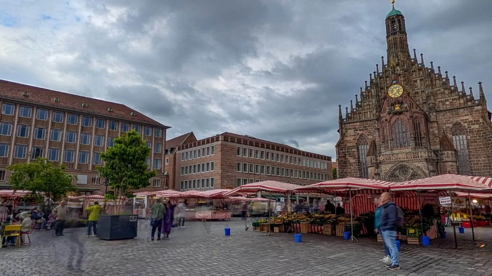 Das bunte Treiben auf dem Nürnberger Hauptmarkt. Vor der historischen Frauenkirche stehen ein mobiler Baum im Pflanzkübel, Marktstände und bunte Stühle.
