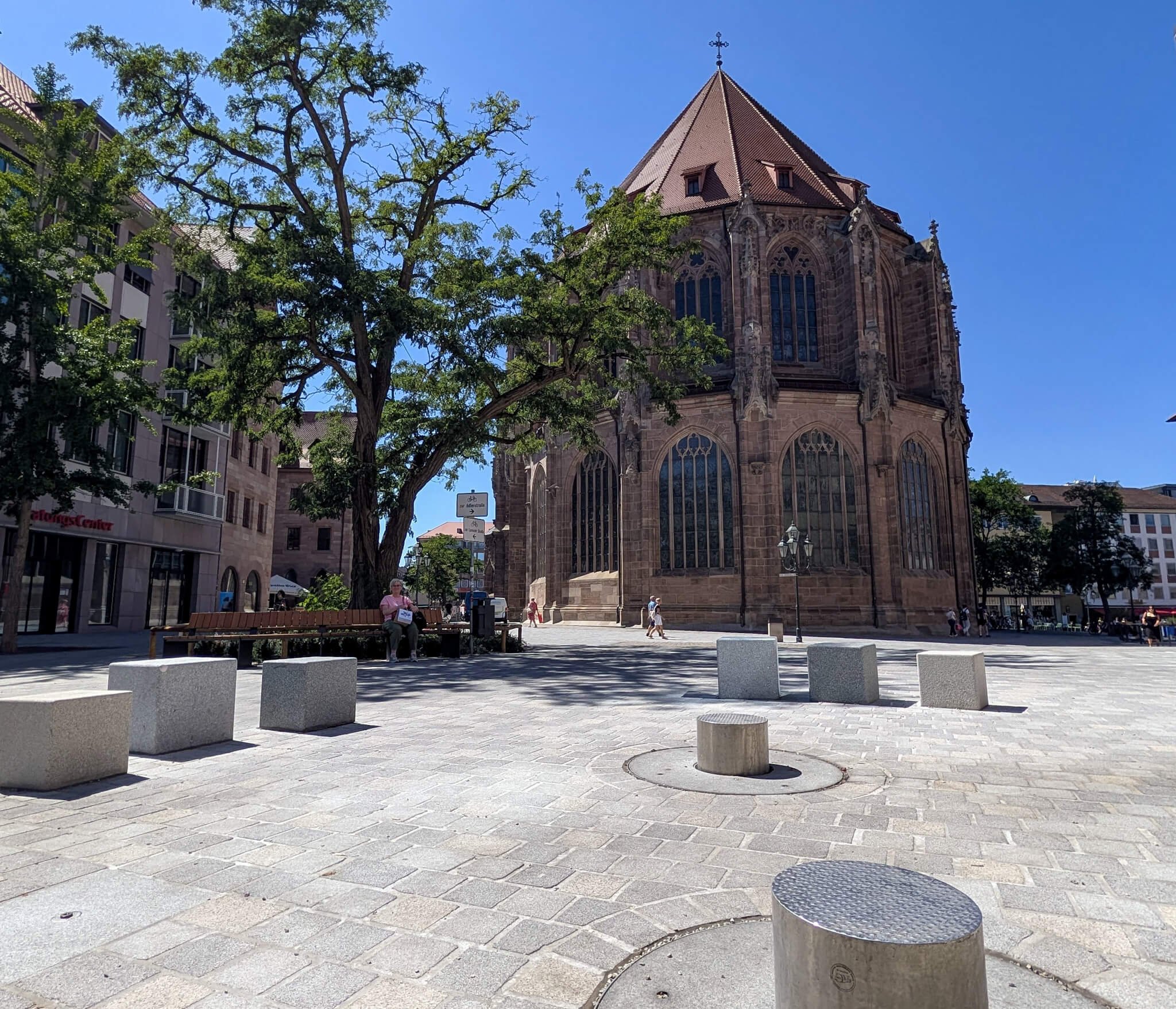 Weite, neu gepflasterte Fläche des Lorenzer Platzes in Nürnberg bei Sonnenschein, mit Betonquadern, einem großen Baum mit Sitzbank im Schatten und dem Chor der Lorenzkirche.
