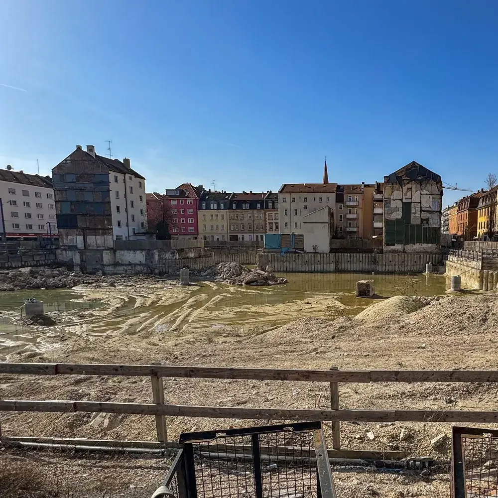 Große, brachliegende Baugrube mit Wasserpfützen am Aufseßplatz in Nürnberg. Im Hintergrund sind städtische Wohnhäuser zu sehen. Auf dem Gelände des ehemaligen Kaufhofs sollen 200 neue Wohnungen entstehen.