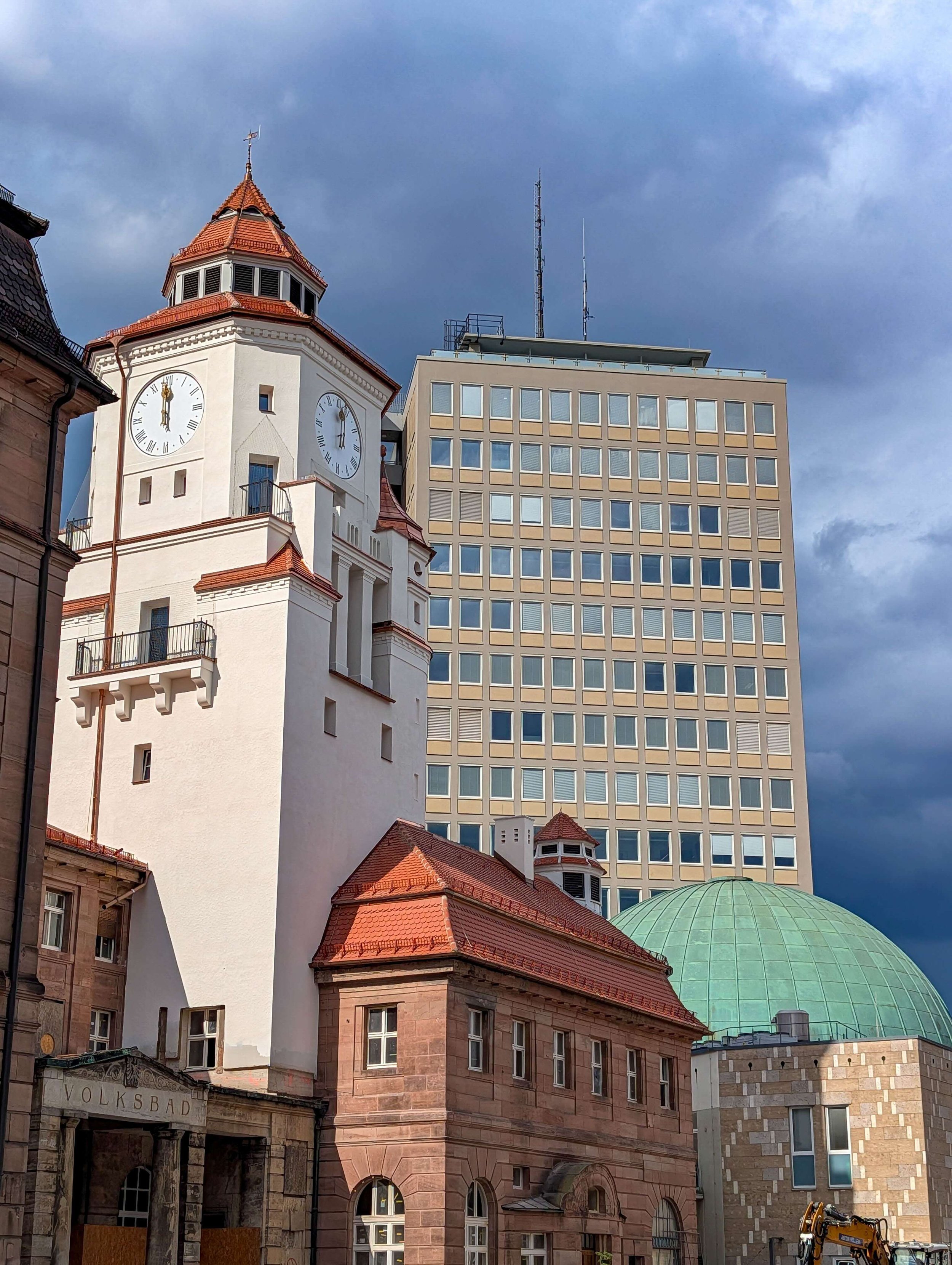 Frisch sanierter Uhrturm des denkmalgeschützten Volksbads Nürnberg neben verwittertem Sandsteinbau, im Hintergrund das Plärrer-Hochhaus und das Planetarium