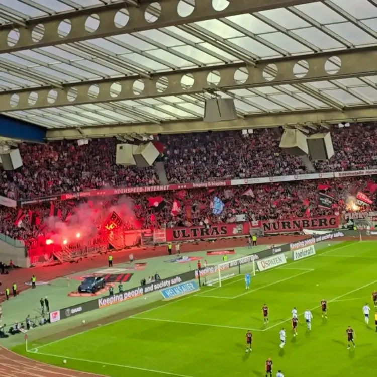 Blick von der Tribüne in das Max-Morlock-Stadion während des Spiels 1. FC Nürnberg gegen Schalke 04 mit roten Rauchtöpfen in der Fankurve und der Beton-Dachkonstruktion.