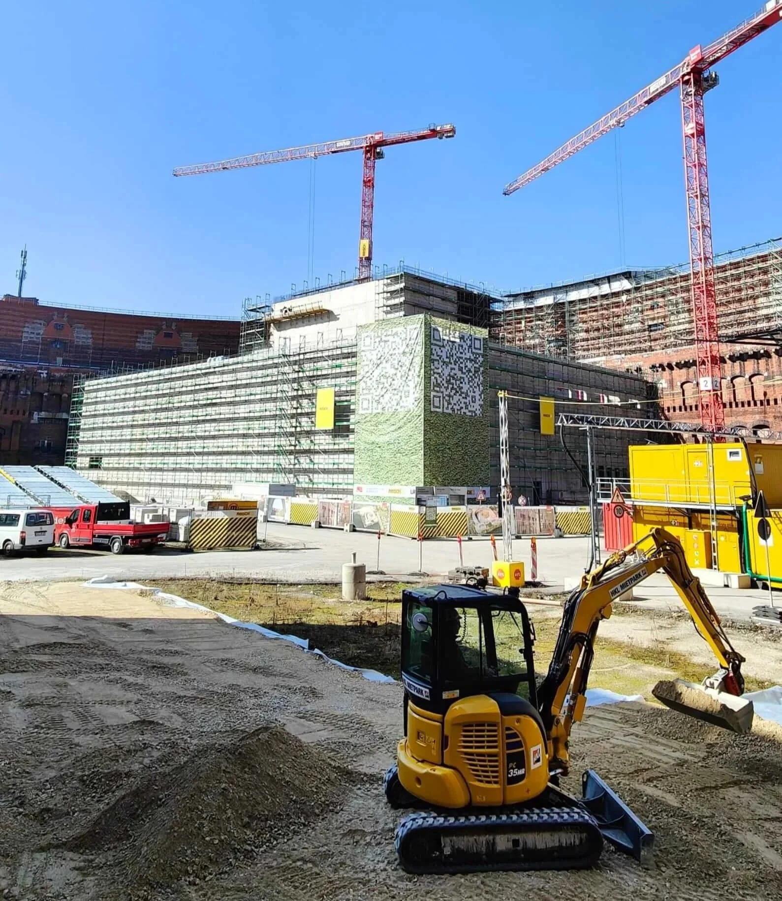 Ein kleiner gelber Minibagger auf einer Baustelle im Vordergrund vor der großflächig eingerüsteten Kongresshalle in Nürnberg unter strahlend blauem Himmel mit zwei großen roten Baukränen