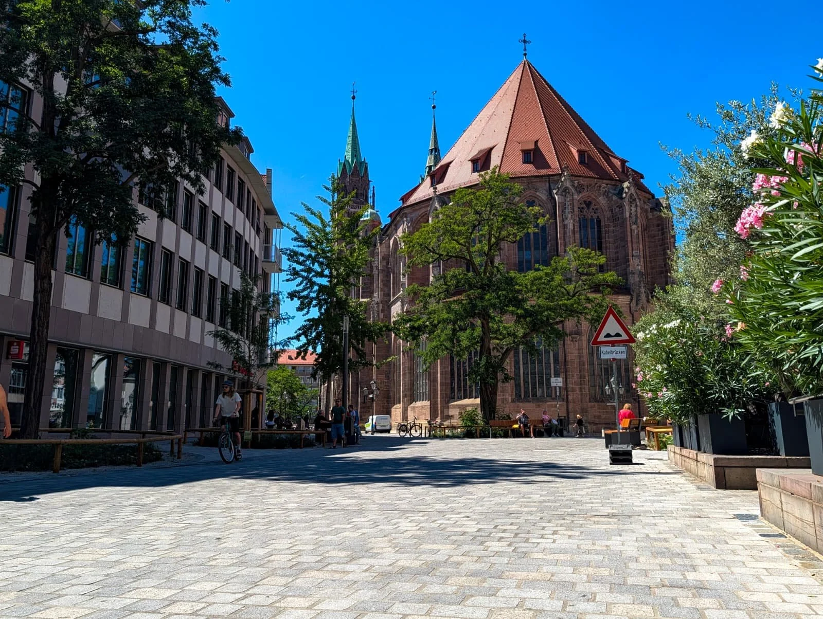 Lorenzer Platz in Nürnberg nach dem Umbau: Blick über das neue  Pflaster zur Lorenzkirche, links Holzkonstruktionen zum Schutz  der Neupflanzungen, rechts blühender Oleander, im Hintergrund  Passanten und ein Radfahrer.