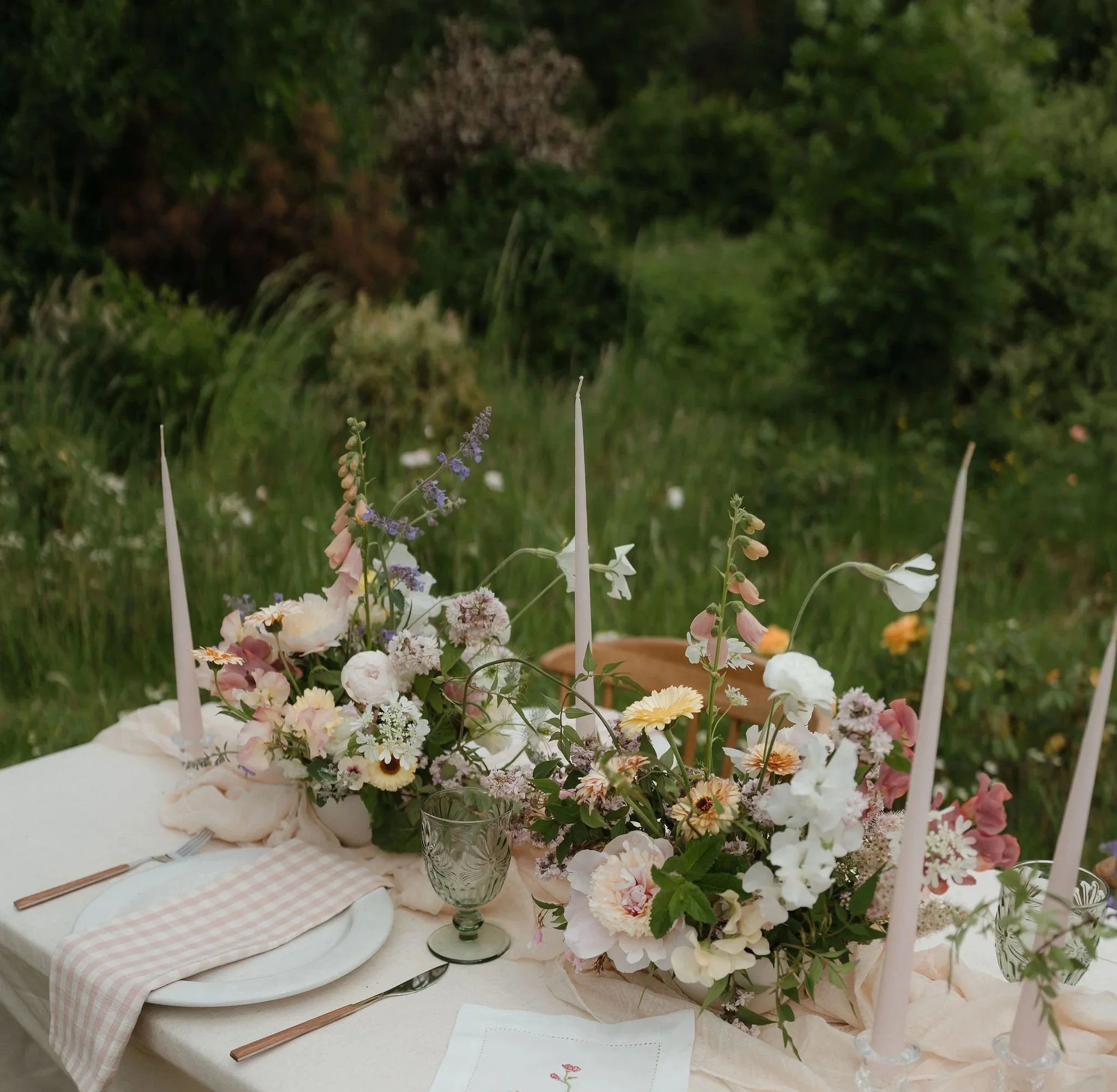 A decorated table with floral arrangements, candles, plates, and utensils set for a special occasion outdoors with greenery in the background.