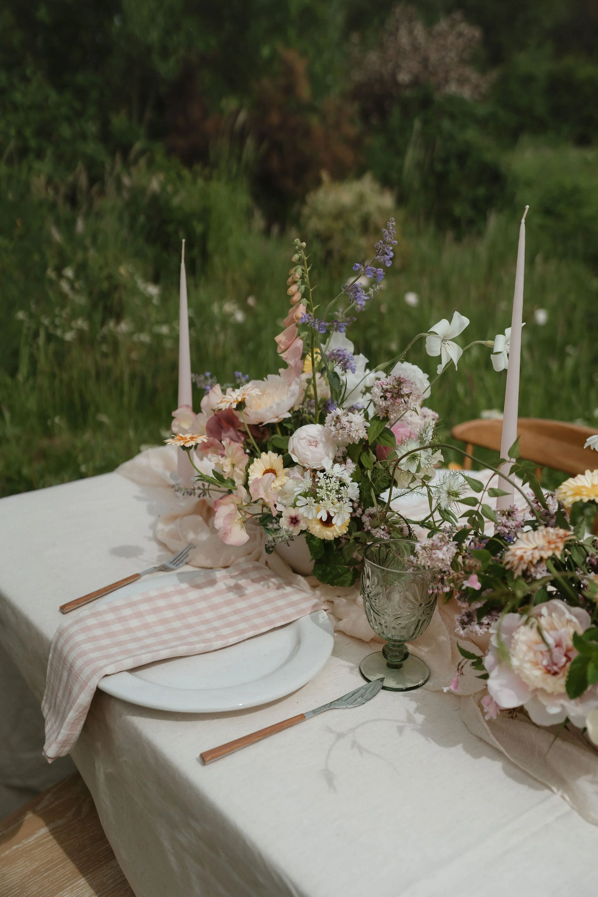 A table set outdoors with a floral centerpiece, pink candles, white plates, silverware, and a pink checkered cloth, with a grassy area and trees in the background.