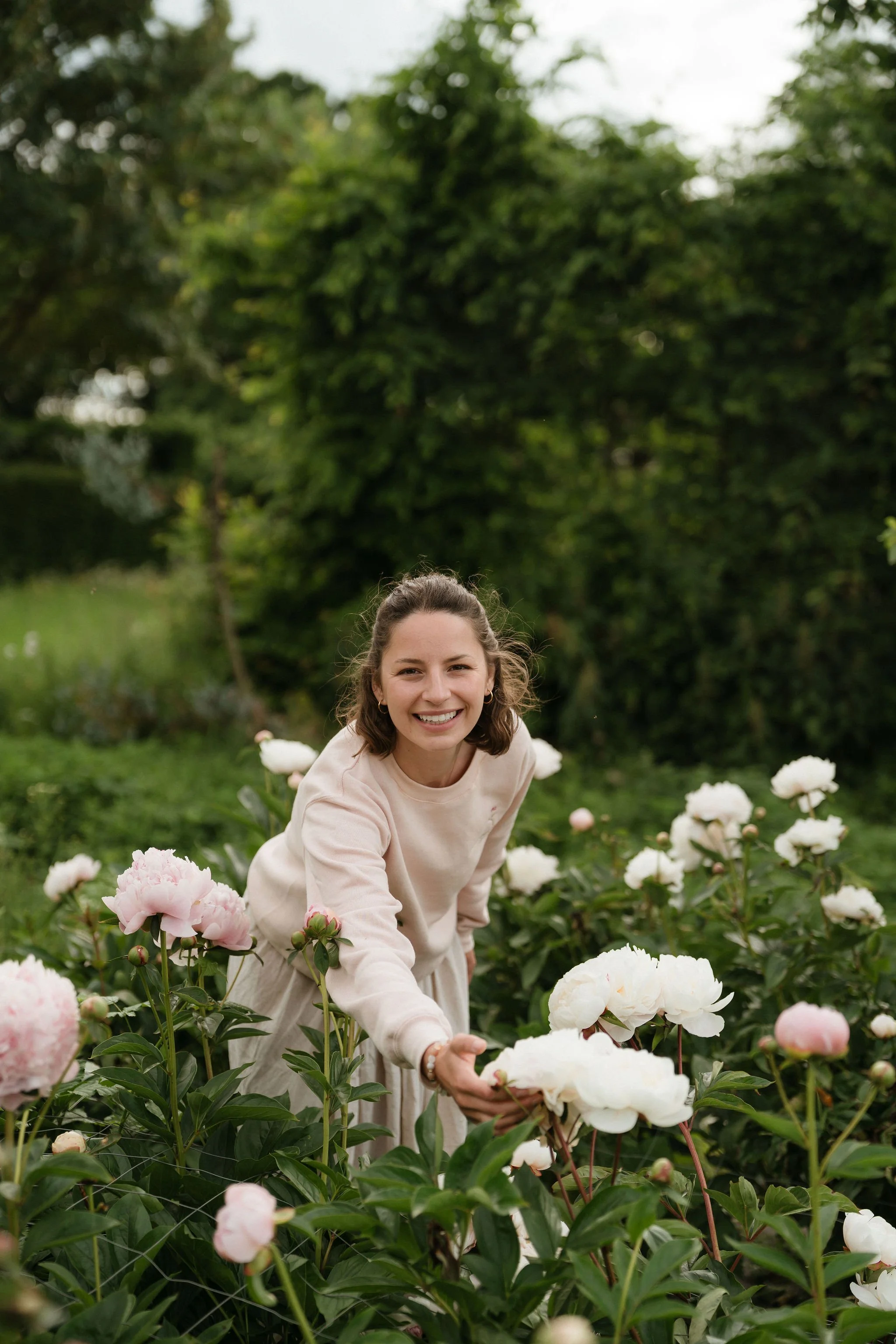 A woman with brown hair in a ponytail, smiling and reaching out to touch white peony flowers in a garden with green trees in the background.