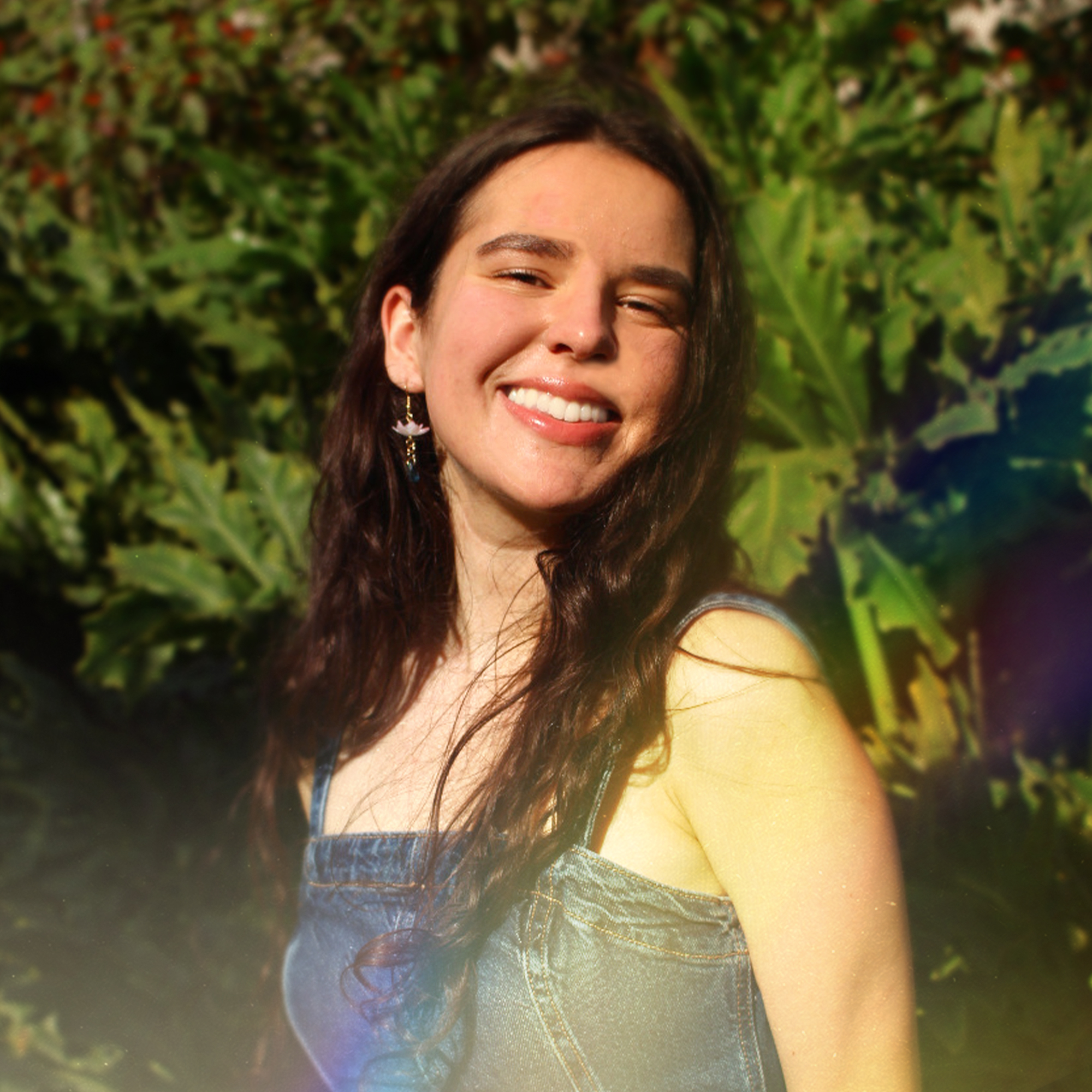 A warm portrait of illustrator and designer Juliet Roselie smiling at the camera, surrounded by lush greenery and layered leaves in a bright, natural setting.