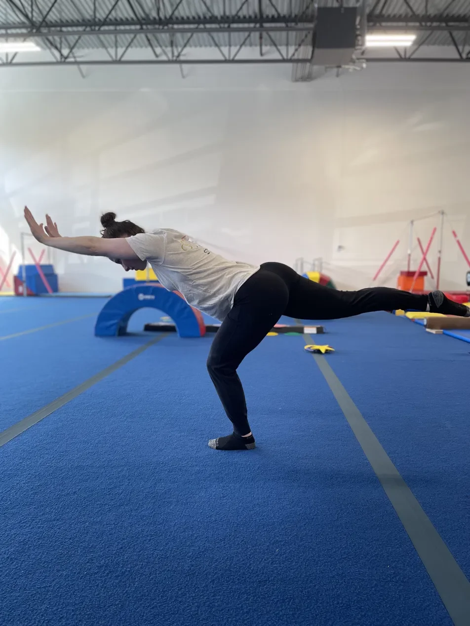 A person in a gymnasium performing a handstand or flip over a padded mat, with gymnastic equipment and colorful foam shapes in the background.