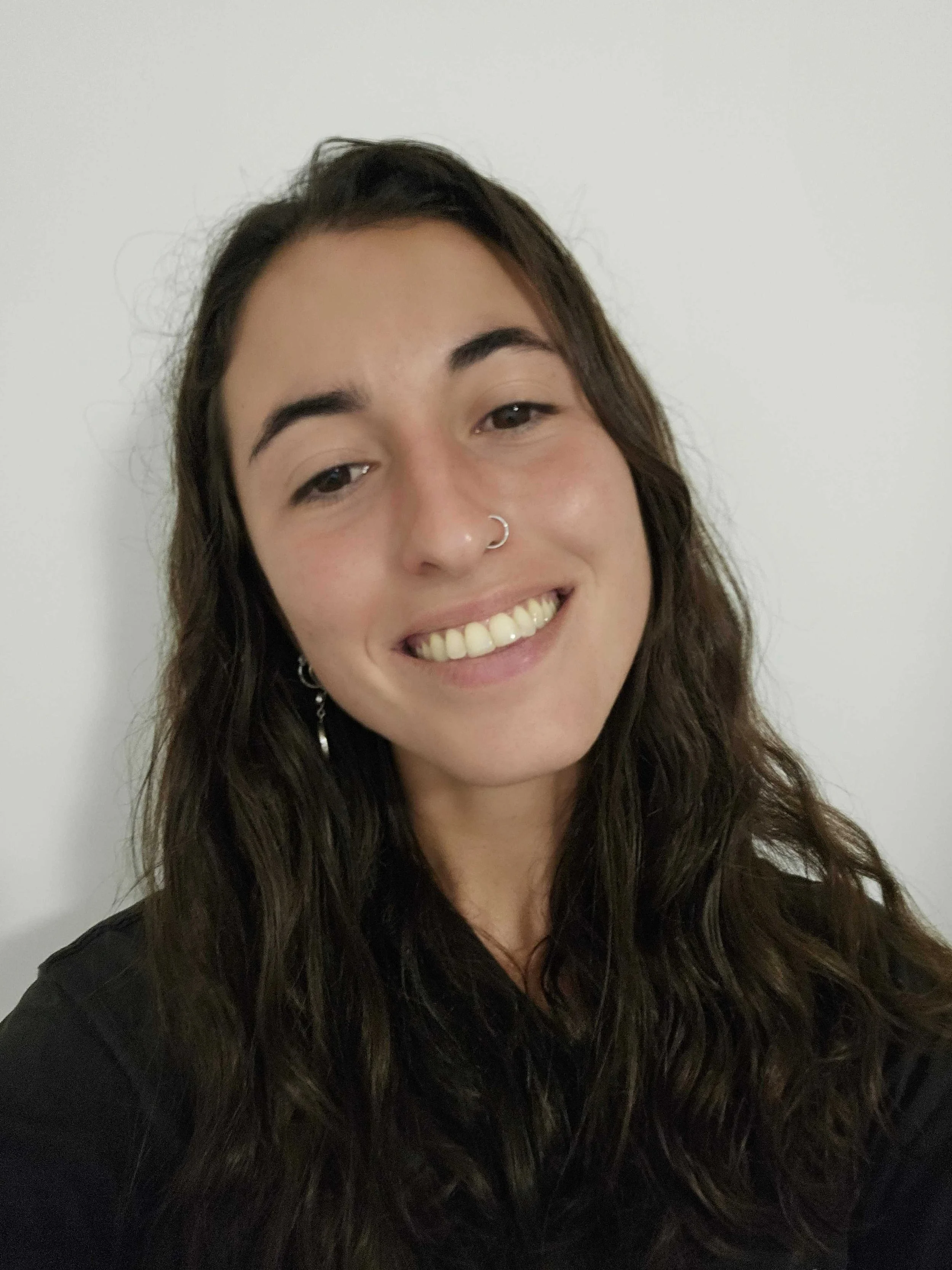 Close-up photo of a smiling woman with long, wavy brown hair, wearing a nose ring and earrings, against a plain white background.