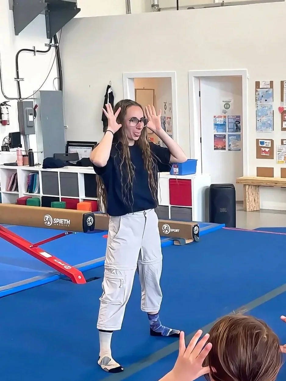 A woman with long hair and glasses standing on a blue gym mat, making a playful face and holding her hands near her head. She is wearing a navy shirt, white cargo pants, and mismatched socks. In the foreground, part of a child's head and hands are visible.