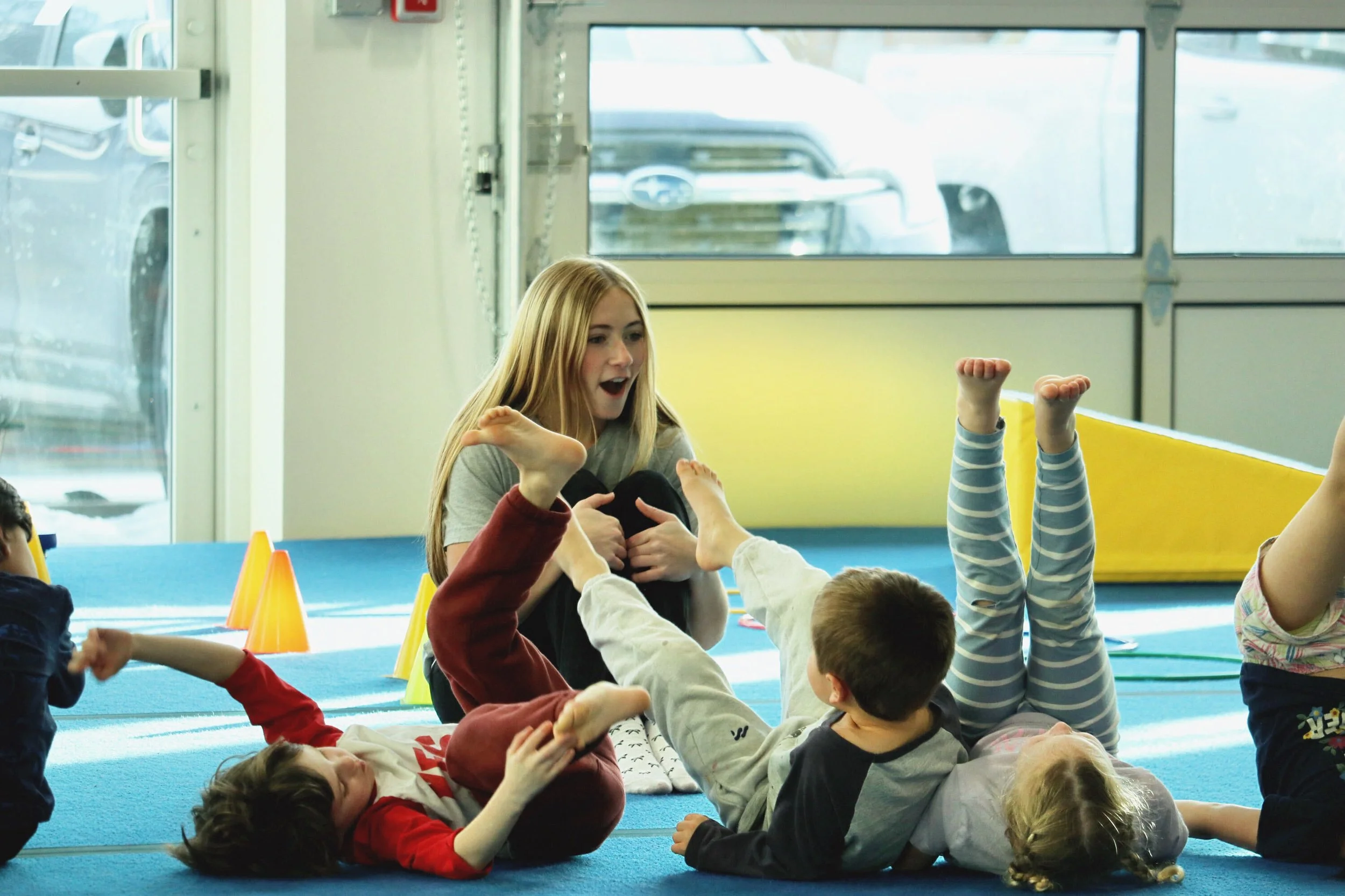 Children lying on a gym floor with orange cones and a woman sitting among them, raising her hand, in a room with large windows and natural light.