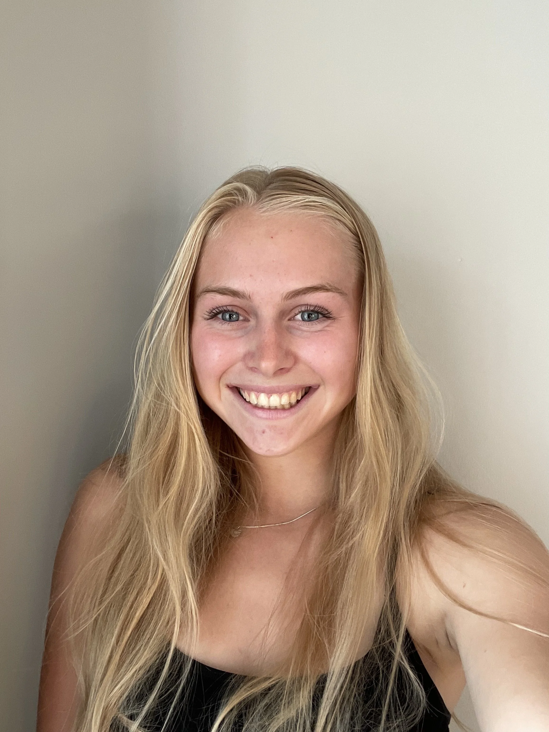 A woman with long, wavy blonde hair smiling at the camera, wearing a black top, against a plain, light-colored wall.
