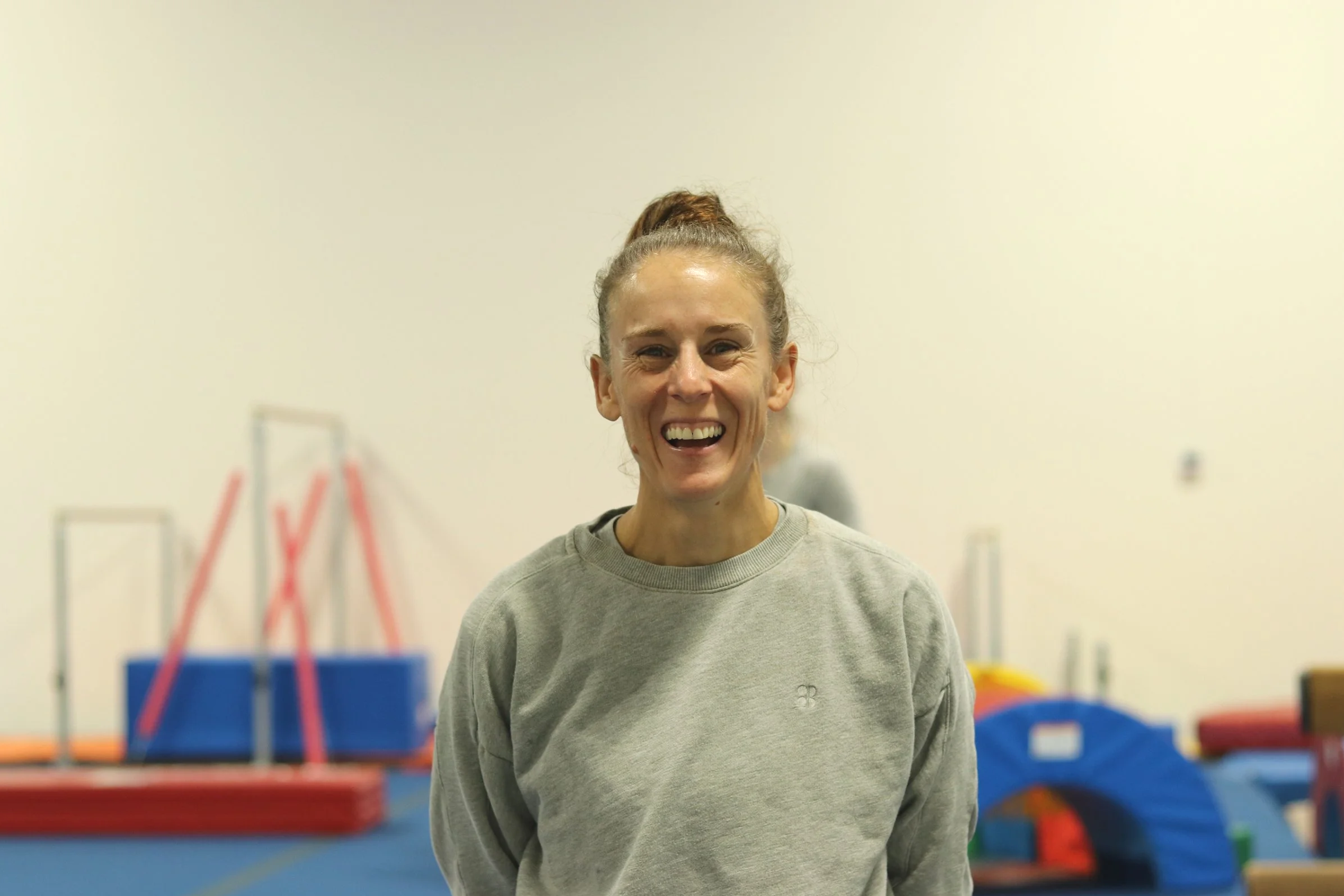 Smiling woman with curly hair tied in a bun, wearing a gray sweatshirt, standing in a gym or sports facility with colorful mats and gym equipment in the background.