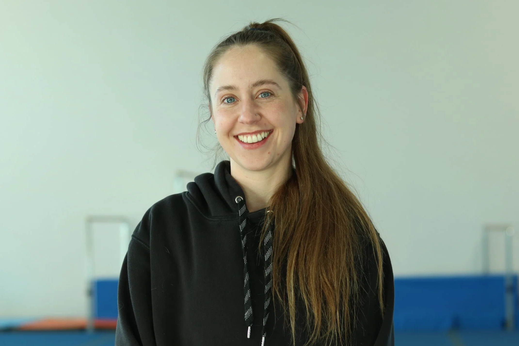A young woman with long brown hair in a ponytail, blue eyes, and earrings, smiling at the camera, wearing a black hoodie, indoors with a blurred background.