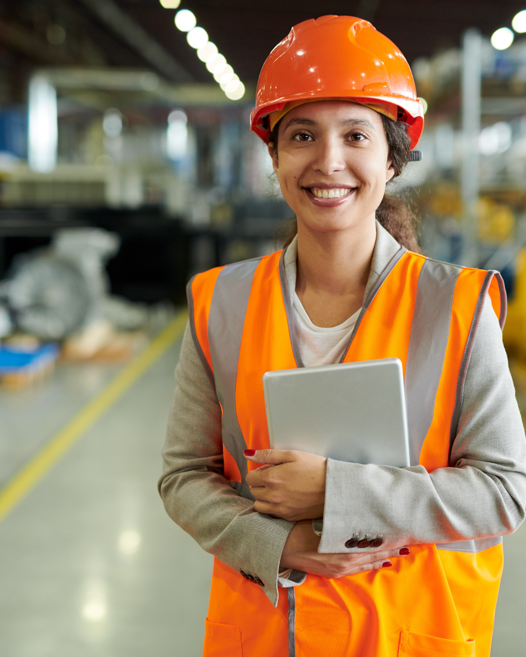 A smiling woman wearing a hard hat and a high-visibility safety vest, holding a tablet, standing inside a warehouse or factory.