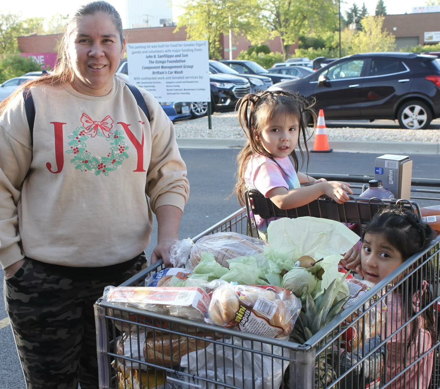 A mother smiling next to a grocery cart filled with fresh food, with her two young children sitting inside the cart at the food pantry.