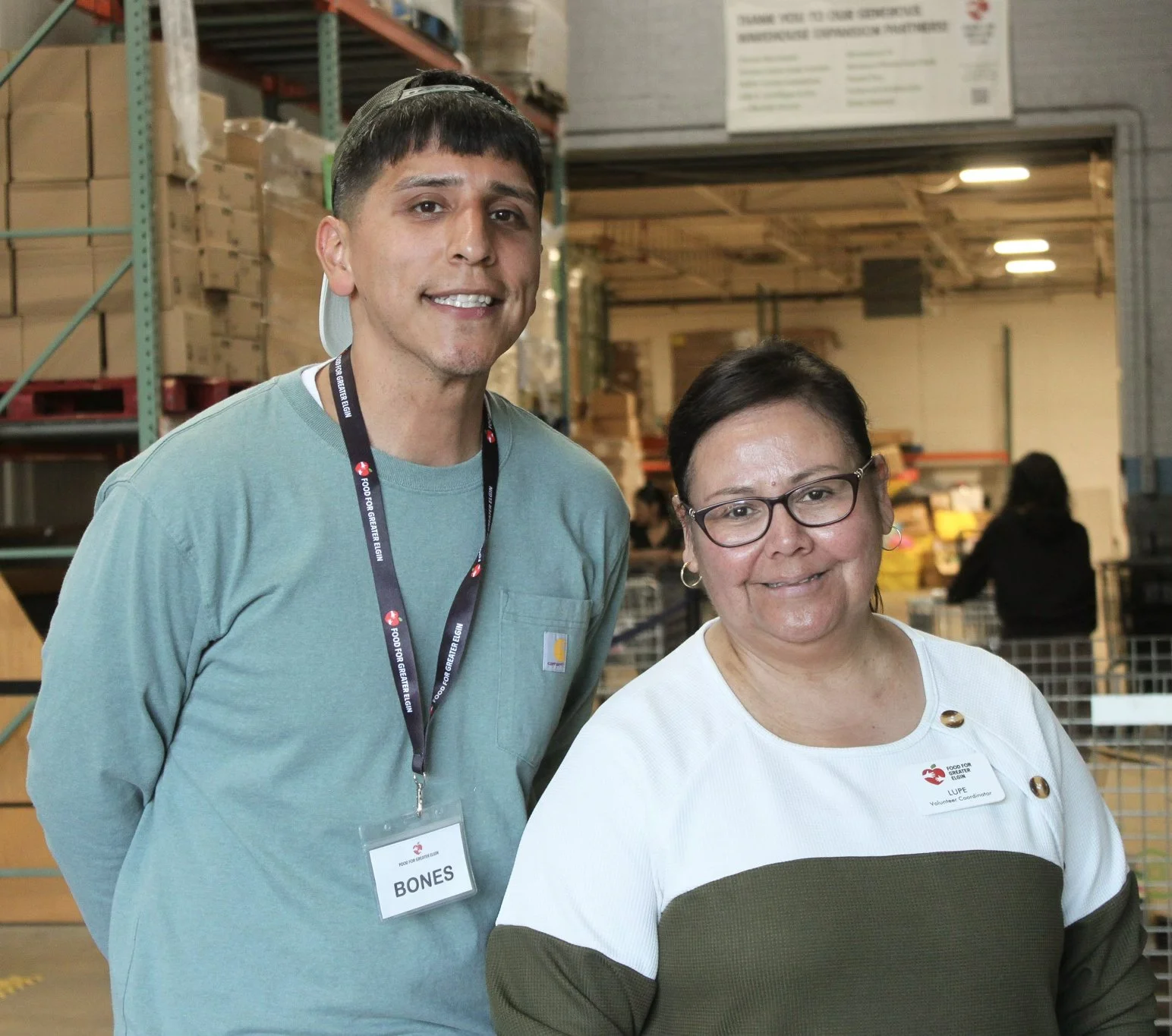Two Food for Greater Elgin employees standing inside the pantry near stocked shelves, with guests shopping in the background.
