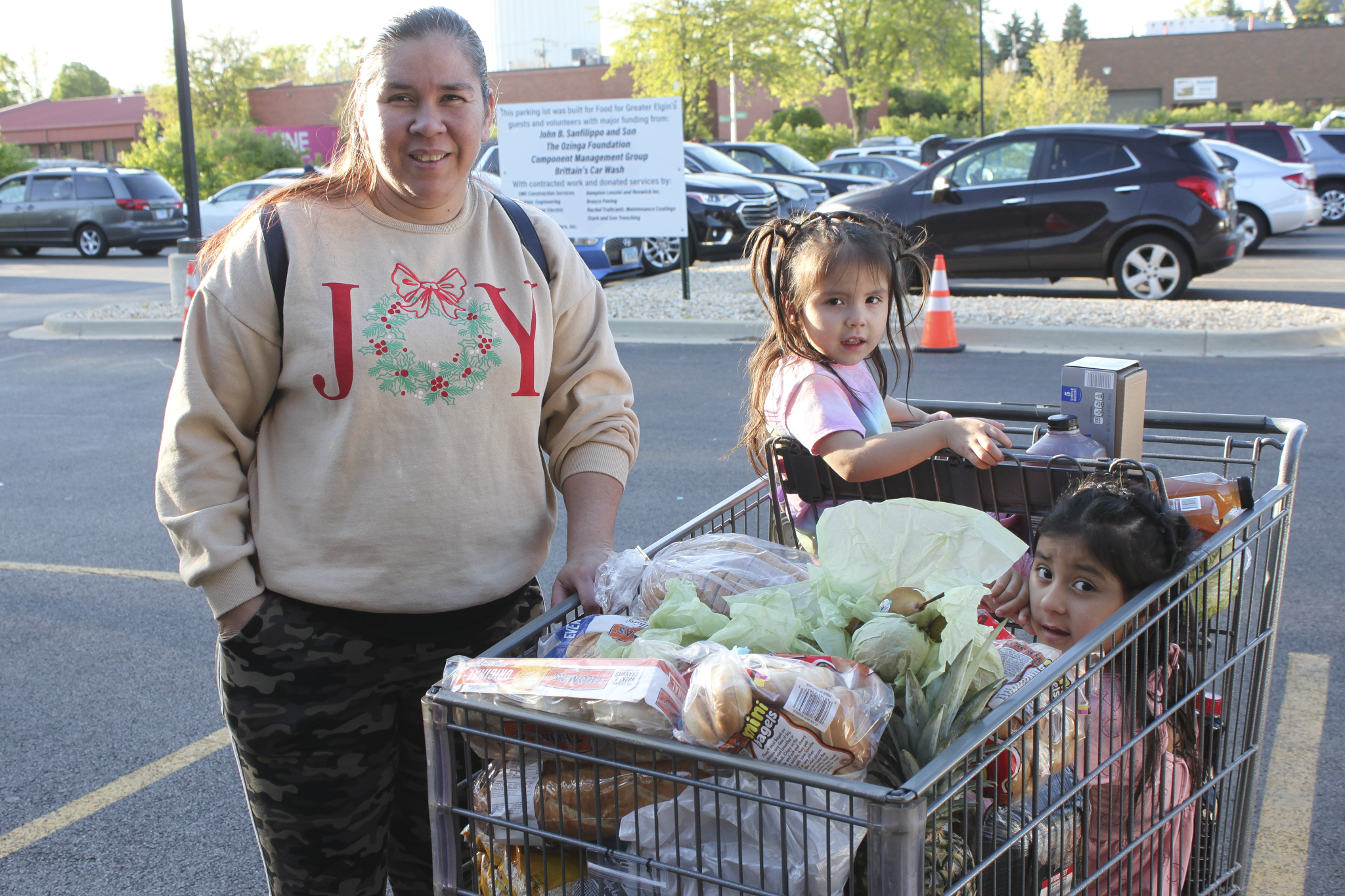 A mom pushing a shopping cart full of groceries with her two young kids sitting inside.