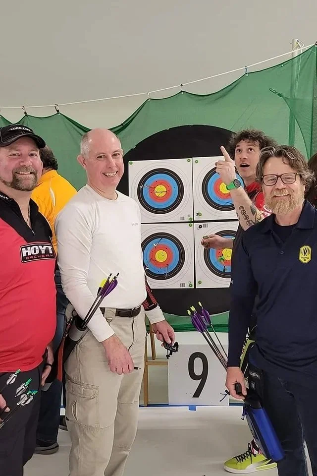 Four men standing in front of archery targets, smiling and holding bows and arrows, at an indoor archery range.
