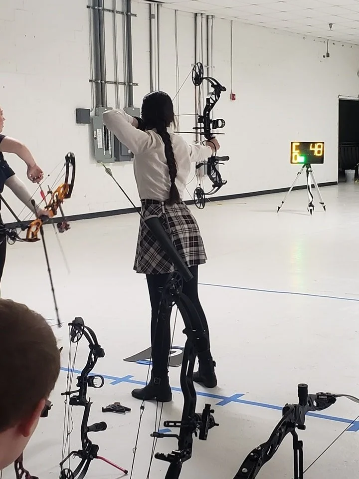 A girl with long dark hair in a plaid skirt and beige top is aiming a bow and arrow in an indoor archery range, with a digital score display in the background.