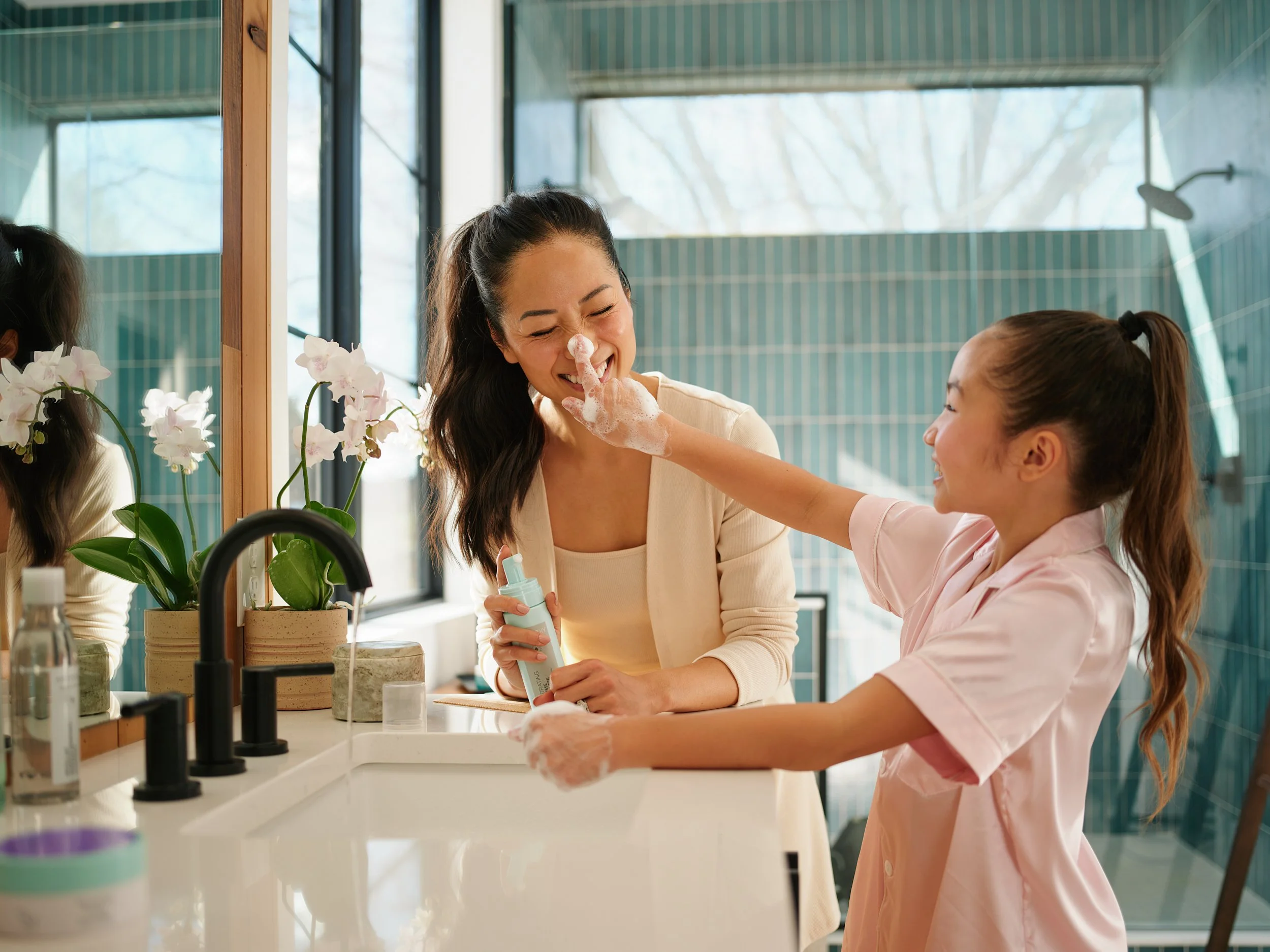 Mother and daughter laughing in a bathroom as the daughter playfully puts soap on her mom’s nose, representing a joyful health and wellbeing moment.
